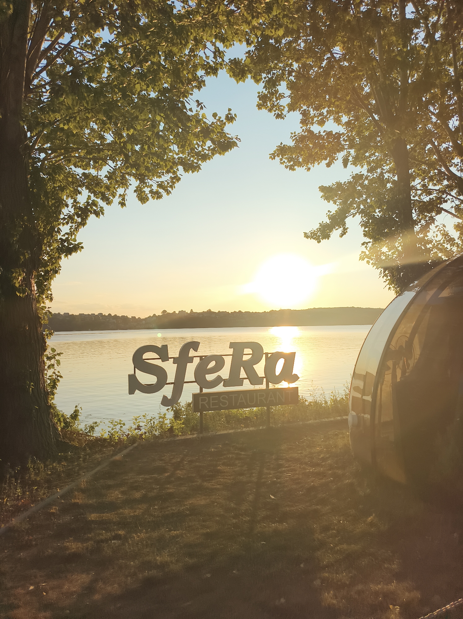 The image depicts a sunset over a body of water, likely the Ternopil Reservoir, near Ternopil, Ukraine. The sun is low on the horizon, casting a warm, golden light across the scene. Trees frame the view, with lush green leaves and dappled sunlight filtering through. In the foreground, a sign reads "Sfera Restaurant" in large, white, three-dimensional letters. To the right, a portion of a dark, reflective, spherical structure, possibly a glamping pod or a unique architectural element, is visible. The water is calm, with subtle ripples reflecting the light. The overall atmosphere is serene and picturesque, suggesting a tranquil evening setting. There are no people visible in the image.