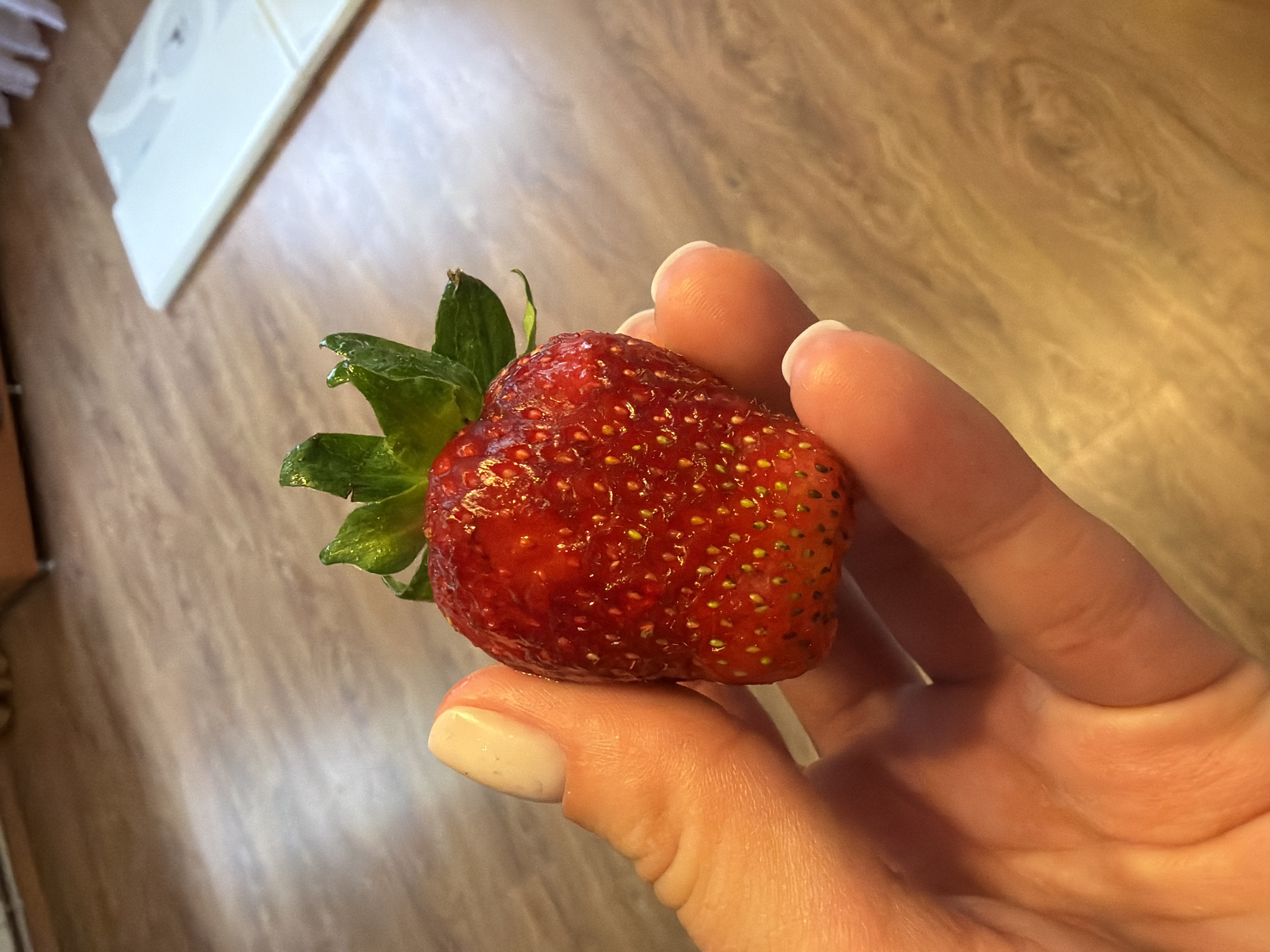 A person's hand holds a single, ripe strawberry. The strawberry is bright red with visible seeds and green leaves at the top. The hand has light skin with a light-colored nail polish on one fingernail. The background is a wooden surface with a slight sheen. A white object, possibly a scale, is visible in the upper right corner. The scene appears to be indoors.