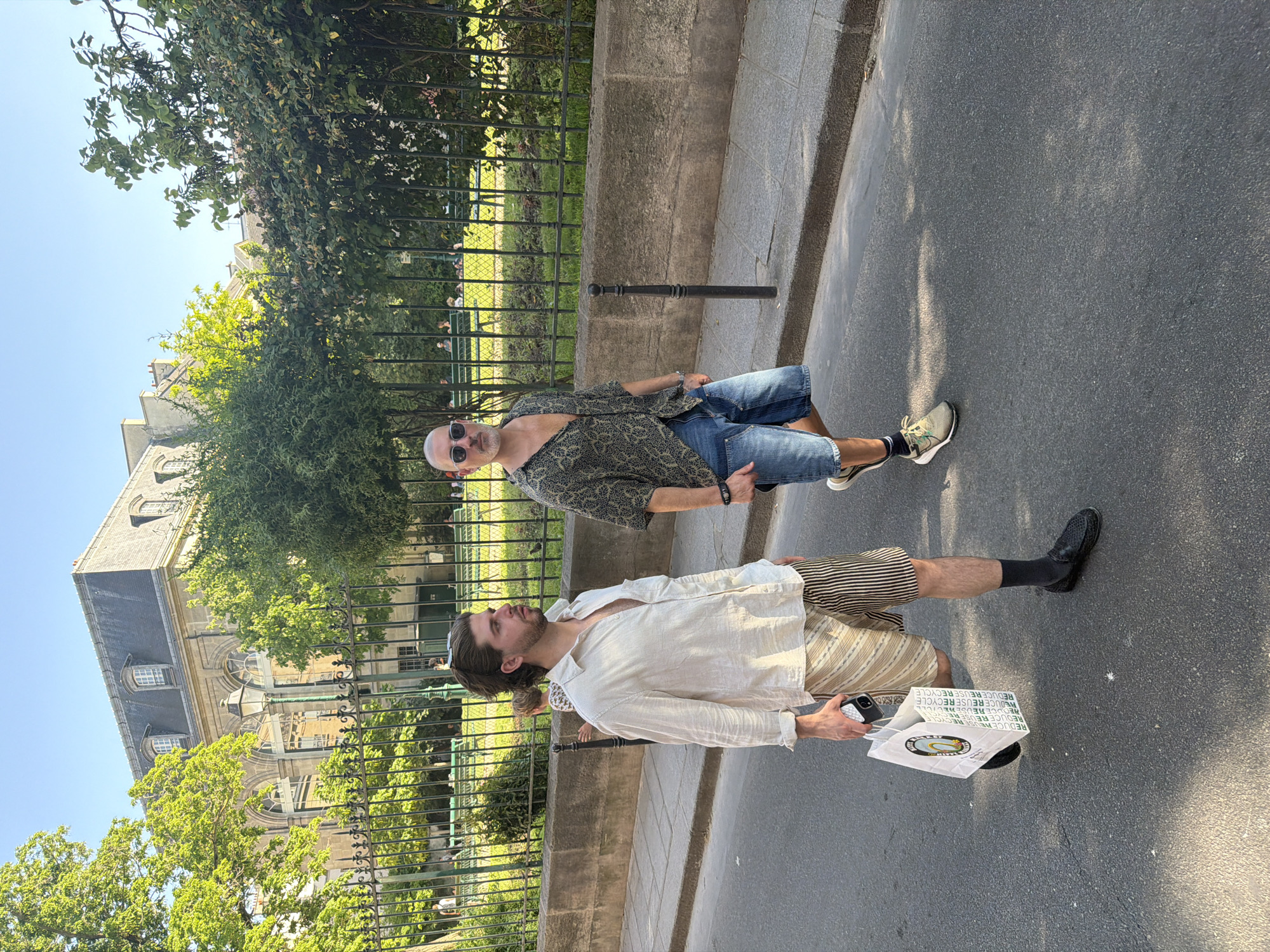 The image captures a street scene in Paris, France, with two men standing on a paved sidewalk next to a low stone wall topped with a black metal fence. Behind the fence is a green space with trees and part of a building visible in the background. The first man is wearing a light-colored linen shirt, striped shorts, black socks, and dress shoes. He holds a mobile phone and a shopping bag with the words "REDUCE REUSE RECYCLE" printed on it. The second man is wearing a dark patterned shirt, denim shorts, sunglasses, and sneakers. The lighting suggests it is daytime.
