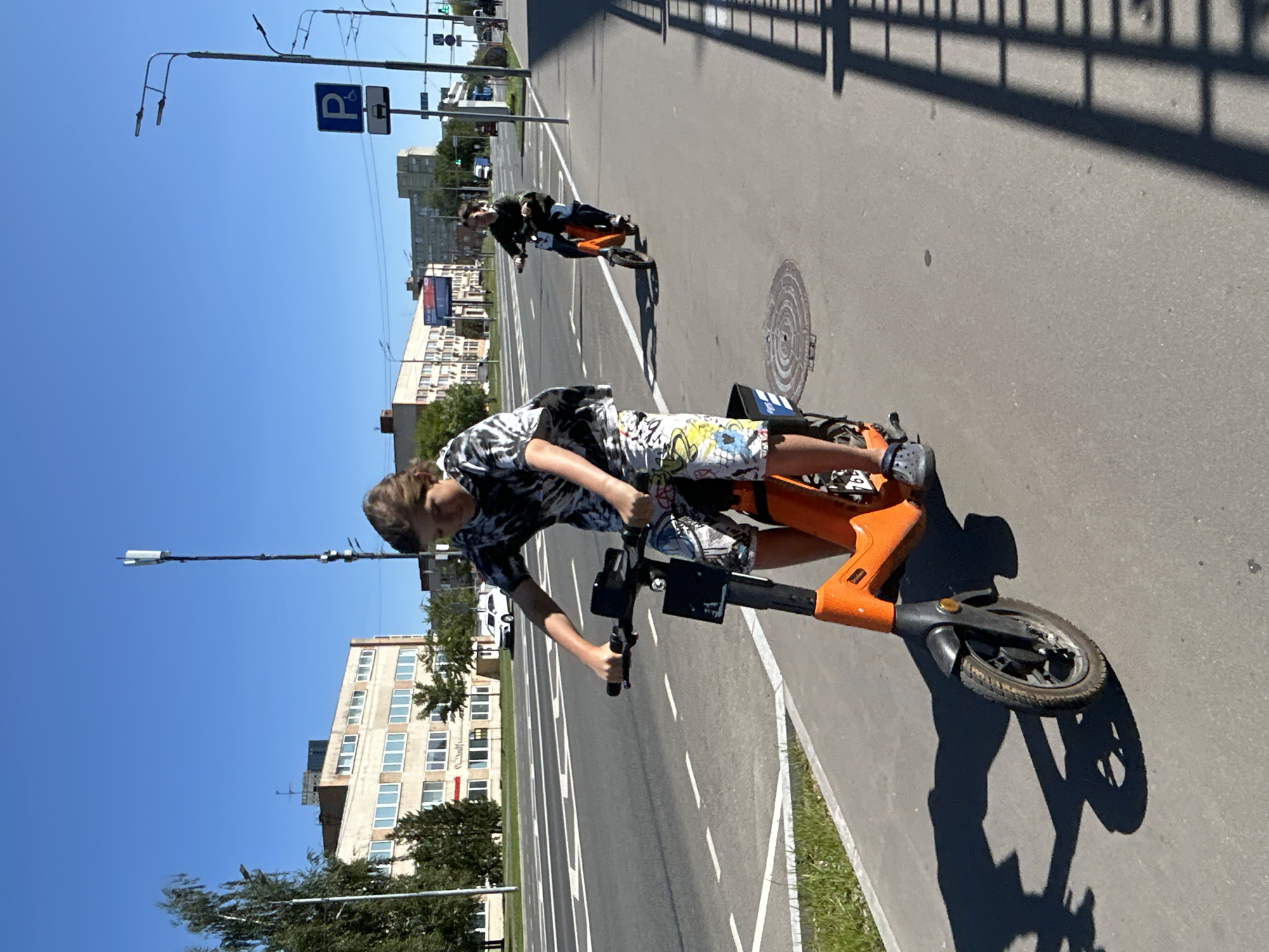 On a sunny day in Moscow, Russia, two young individuals are seen riding electric scooters on a paved sidewalk adjacent to a multi-lane road. The person in the foreground, wearing a black and white tie-dye t-shirt and patterned shorts, is actively riding an orange electric scooter. Their hands are on the handlebars, and their feet are on the footrest. Behind them, a second person on a similar orange electric scooter is also riding along the sidewalk. The environment includes buildings with multiple windows and a clear blue sky, characteristic of an urban setting. Streetlights and traffic signs, including a parking sign and a road sign, are visible along the roadside. The road itself has lane markings, and a white car is observed driving on it. The sidewalk is bordered by a grassy verge. The image captures a moment of recreational activity in a city environment.
