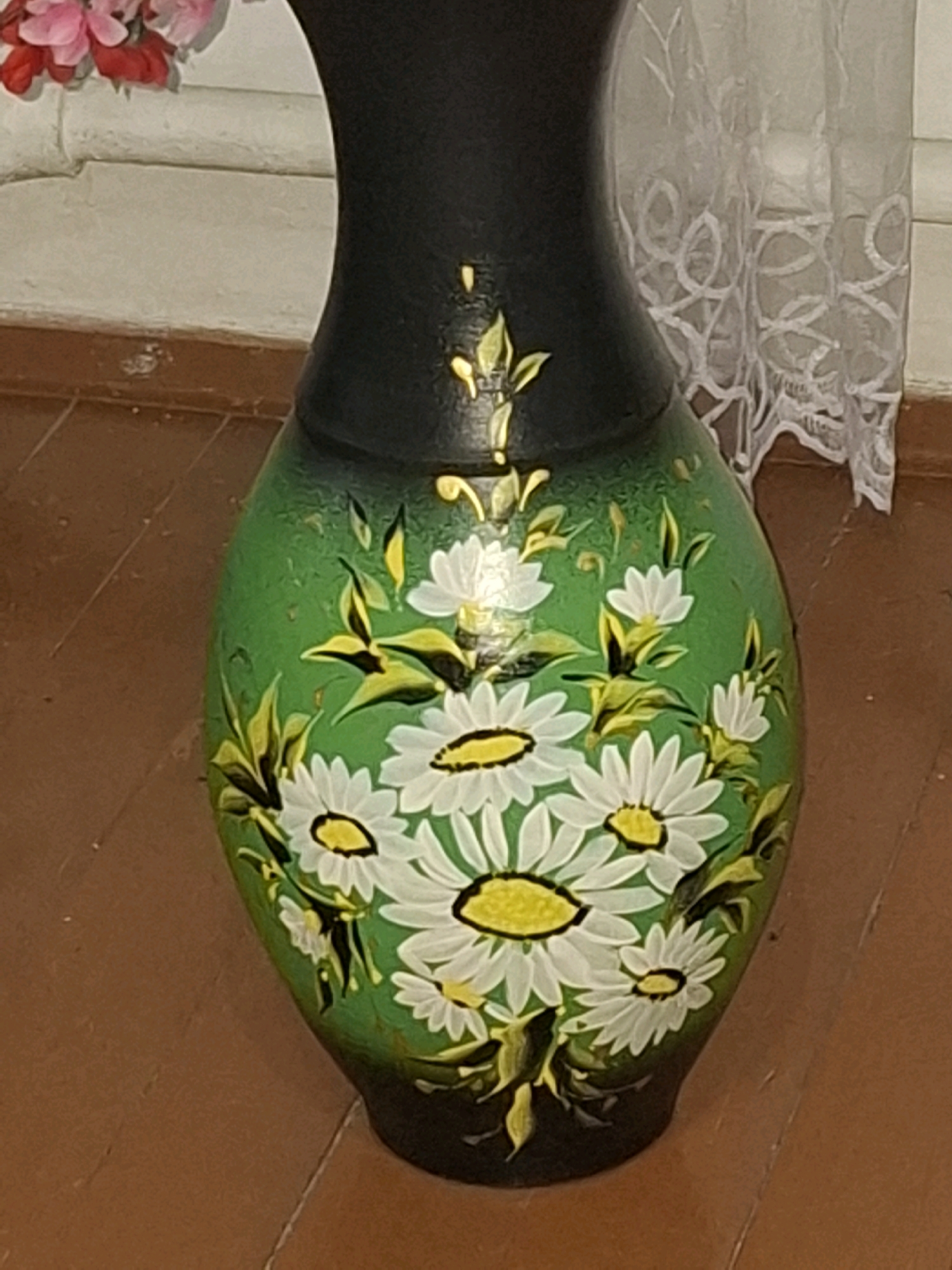A close-up, medium shot shows a decorative vase with a black neck and a green body painted with white daisies and green leaves. The vase is placed on a brown wooden floor. In the background, a white brick wall is visible, with pink flowers partially in frame at the top left, and a white lace curtain on the right. The scene appears to be indoors, likely in a residential setting.