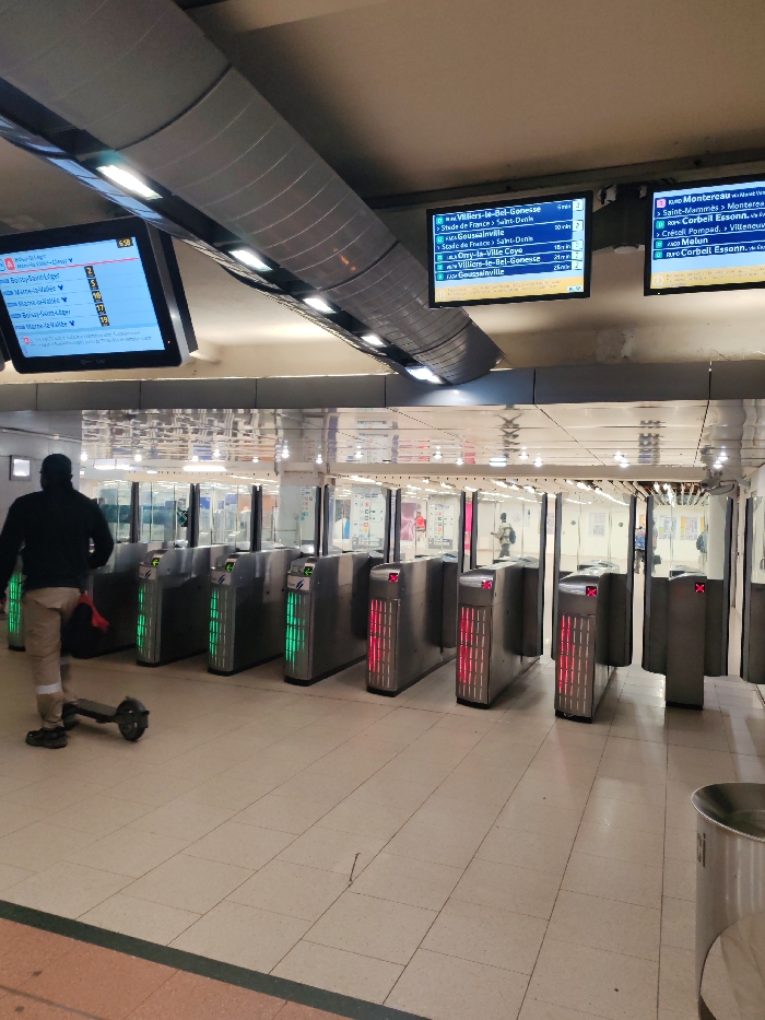 This image depicts the interior of a public transit station in Neuilly-Plaisance, France, focusing on a row of automated ticket barriers. In the foreground-left, a male individual, viewed from behind, wears a dark jacket and light-colored trousers, standing adjacent to the barriers. An electric scooter rests horizontally on the light-colored, square-tiled floor near his feet.
Seven ticket barriers are visible across the mid-ground. The three leftmost barriers display vertical green illuminated panels, indicating they are open for passage. The four barriers to their right each display a red 'X' symbol, indicating they are closed or inactive.
Above the barriers, three large, black-framed digital display screens are mounted. The leftmost screen, partially obscured, shows French train destinations including "BOISSY-SAINT-LEGER" and "MARNE-LA-VALLEE-CHESSY." The central screen displays RER B line destinations and times, such as "Villiers-le-Bel-Gonesse," "Stade de France > St-Denis," and "Goussainville." The rightmost screen displays RER D line destinations and times, including "Montereau" and "Corbeil Essonne."
The ceiling features a prominent, curved metallic duct system with integrated linear lighting fixtures, providing bright overhead illumination. The floor is tiled with light-toned ceramic squares. A thin, dark object, resembling a nail, is visible on the floor in the mid-ground. A cylindrical stainless steel waste bin is partially visible on the far right of the frame. The background beyond the barriers is a series of glass panels and a brightly lit passage with indistinct figures.