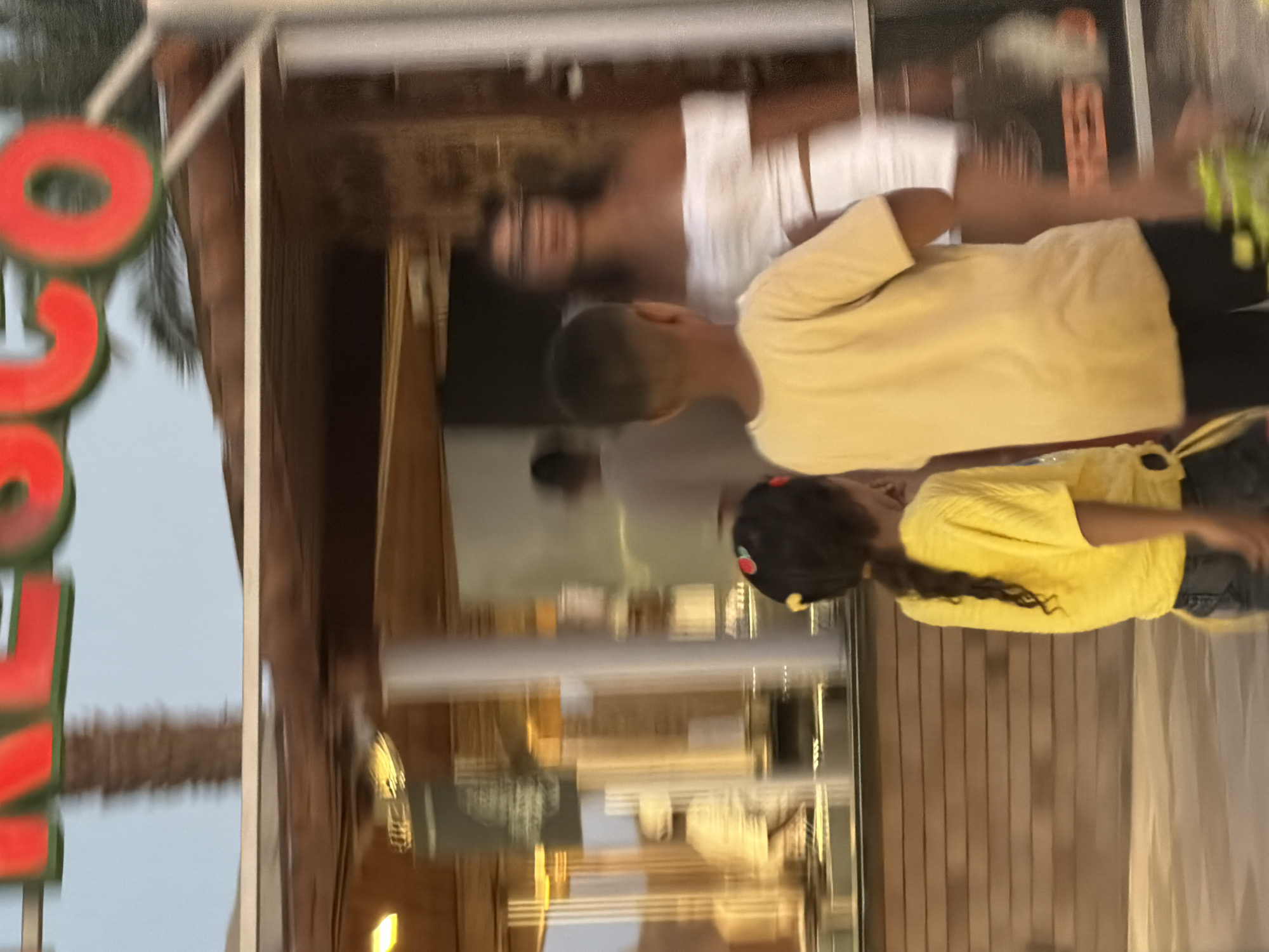 In Sharm El Sheikh, Egypt, at a location featuring a prominent red sign with green lettering that reads "FRESCO," two children, a boy and a girl, stand facing away from the camera. The boy wears a yellow t-shirt and dark pants, while the girl wears a yellow shirt and dark pants, with a hair accessory on her head. Behind them, a woman wearing a white off-the-shoulder top and a white skirt is visible, appearing somewhat blurred. The background suggests an establishment with a glass facade, possibly a restaurant or shop, with internal lighting and wooden paneling. The overall scene is captured in motion, indicating it was taken from a moving vehicle or the subjects are in motion, resulting in a slight blur across the image.