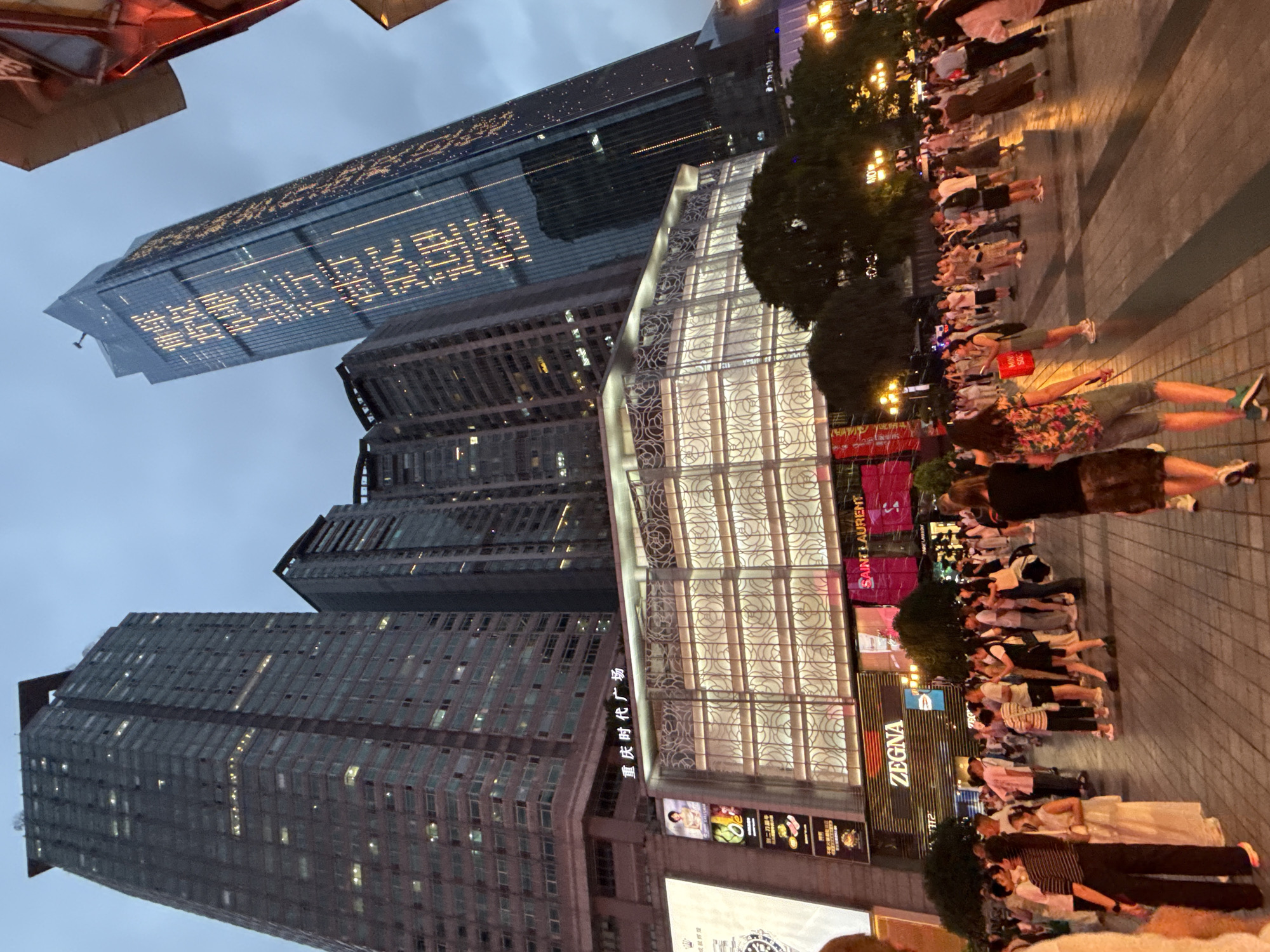 The image depicts a bustling street scene in Yuzhong District, China, at dusk. Towering skyscrapers dominate the background, with one particularly tall, glass-fronted building illuminated by what appears to be a large, vertically displayed message written in lights. In the foreground, a crowd of people is gathered on a paved plaza. Several retail establishments are visible, including one with a large "ZEGNA" sign. The overall atmosphere is one of urban activity and commercial vibrancy.