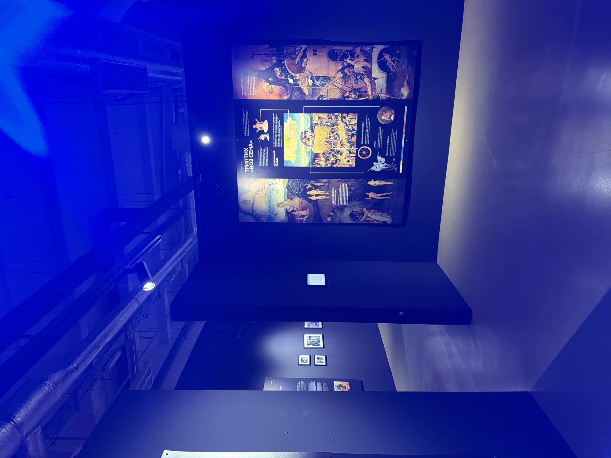 This media file shows an exhibit in a dimly lit room, bathed in blue light. The primary subject is a large, vertically oriented display featuring a triptych painting, likely a reproduction. The triptych is divided into three panels, each depicting a scene with multiple human figures. The style appears to be historical or religious art, with detailed, crowded compositions. Text is visible on the display, written in a language that appears to be Cyrillic.
Below the main triptych, a series of smaller framed images are mounted on a dark wall. These images are less clear but seem to be photographs or prints. Further down, another display panel with text and images is visible. The overall setting suggests a museum or art gallery, with industrial elements like pipes and lighting fixtures visible in the background, particularly in the upper left portion of the image, which is heavily influenced by the blue lighting. The atmosphere is somber and focused on the artwork. There are no clear indicators of time of day or weather, and the emotions conveyed by the artwork are difficult to ascertain from this perspective, though the subject matter of the triptych may suggest dramatic or moral themes. The blue lighting creates a somewhat artificial and dramatic ambiance.