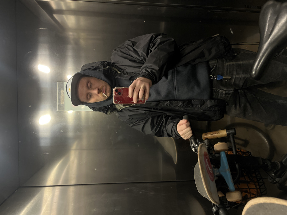 A male individual is depicted in a mirror selfie inside an elevator. He is Caucasian, wearing a dark baseball cap with a light brim, a dark hooded jacket over a dark hooded sweatshirt, and dark faded jeans. A light-colored object, consistent with a hand-rolled cigarette or small cigarillo, is positioned in his mouth. He holds a red-cased smartphone in his right hand to capture the image. A silver ring is visible on his left ring finger, which grips the handlebar of a bicycle. A keychain with a blue plastic key and a metal key hangs from his jeans. A skateboard with a light-colored deck, dark trucks, and light blue bushings is mounted horizontally on the front of the bicycle, above a dark basket. The elevator interior features brushed metallic walls displaying scratches and reflections of overhead artificial lighting. The image contains no visual information to confirm the specific location within Tel-Aviv, Israel.