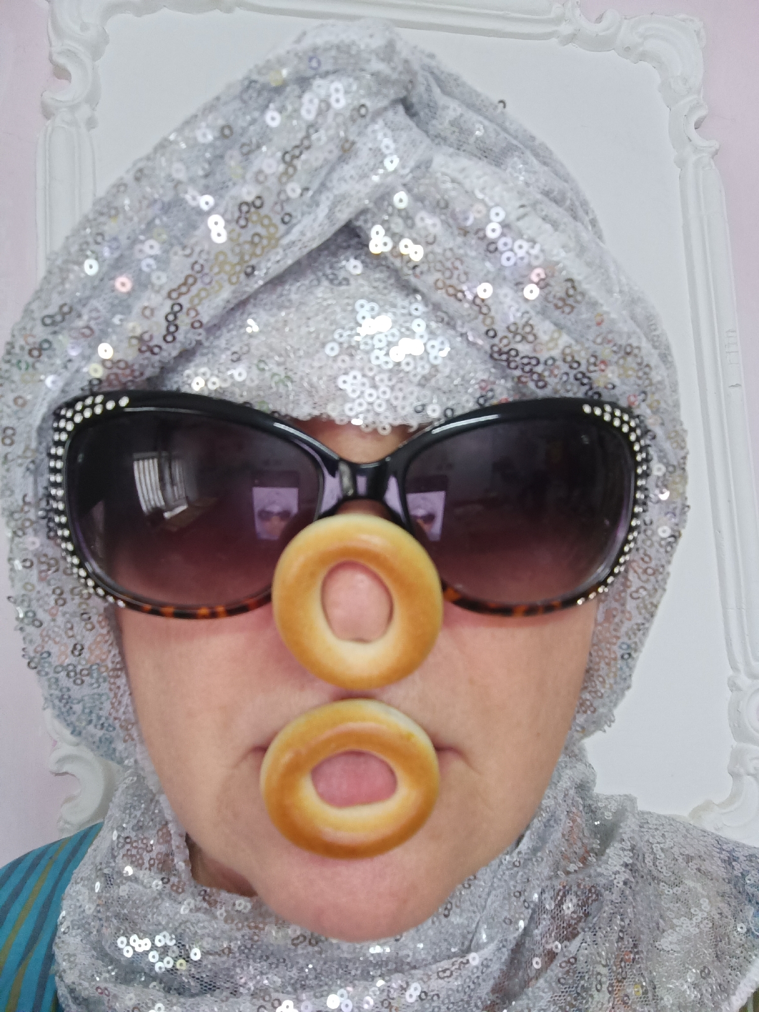 In Tyshky, Ukraine, a person is humorously posing with two small bread rings positioned to resemble a nose. They are wearing a silver sequined turban and large, dark sunglasses adorned with small white rhinestones. The background features a white ornate frame against a pale pink wall.