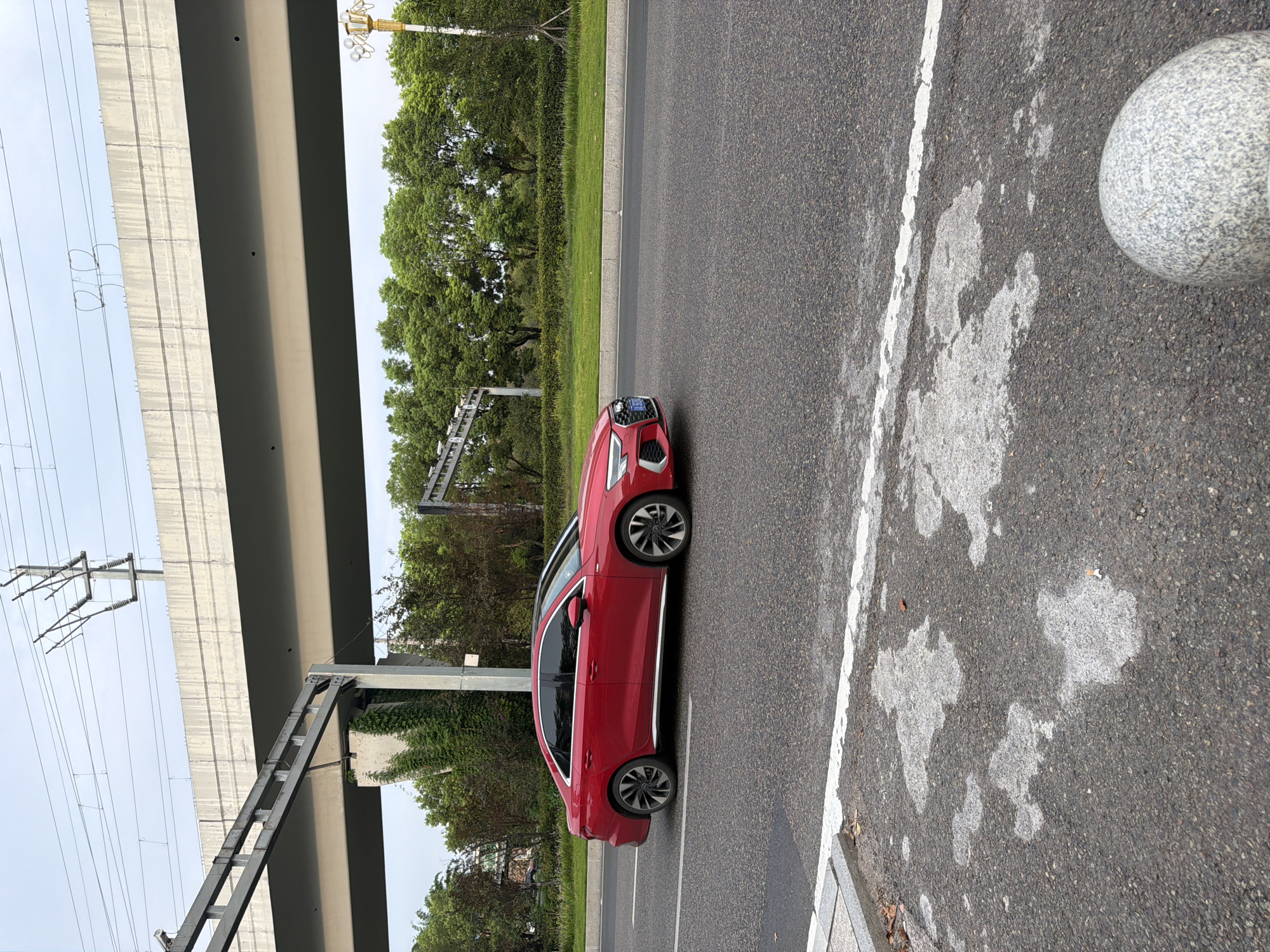 A red Audi sedan drives on a paved road under an elevated highway structure in Jiangjin, China. The car, viewed from the side, is traveling from left to right. The elevated highway is a grey concrete structure supported by pillars, with electrical wires running above it. Lush green trees and vegetation line the opposite side of the road. The road surface is asphalt, with white lane markings visible. The foreground shows a grey concrete curb and a spherical stone bollard. The sky is overcast.