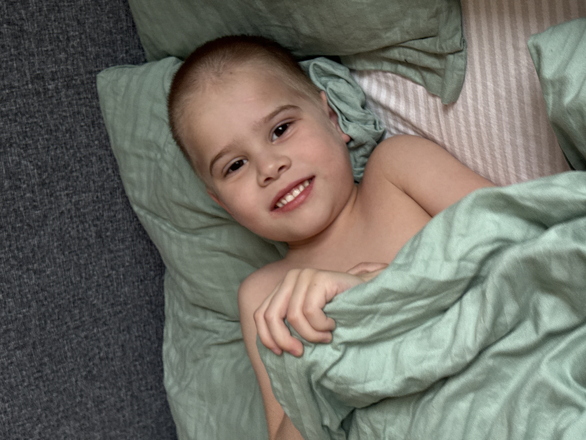 A young boy with a shaved head lies in a bed with green sheets and pillows. He is partially covered by the green sheet and is smiling. The bed has a striped pattern. The bed is next to a gray surface, possibly a couch or the floor. There is no discernible information to determine that the location is in the city of Chaikovsky, Russia.
