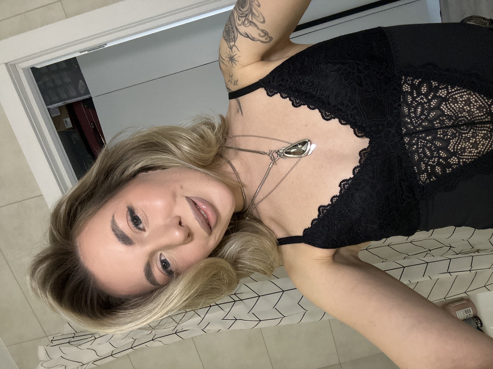 A fair-skinned female subject, estimated to be in her late 20s to early 30s, with blonde, shoulder-length hair, is depicted in a self-portrait. She is smiling faintly while looking directly at the camera. The subject wears a black lace camisole or bodysuit. Two silver necklaces are visible: a choker and a longer chain featuring a large, dark, teardrop-shaped gemstone pendant. Tattoos are present on her left arm and shoulder, including a celestial motif with stars and an abstract design on the upper arm. Her right arm is extended, holding the camera out of frame.
The environment is an indoor setting, likely a residential space. To the left, a white shower curtain with a black geometric pattern (interlocking lines) is visible, with light-colored tiled walls behind it and partially visible bottles on a surface below. To the right, a white door frame opens into another interior space or closet containing various stored items, including cardboard boxes and a red object. The specific city of Khimki, Russia, cannot be determined from the visual evidence within the image.