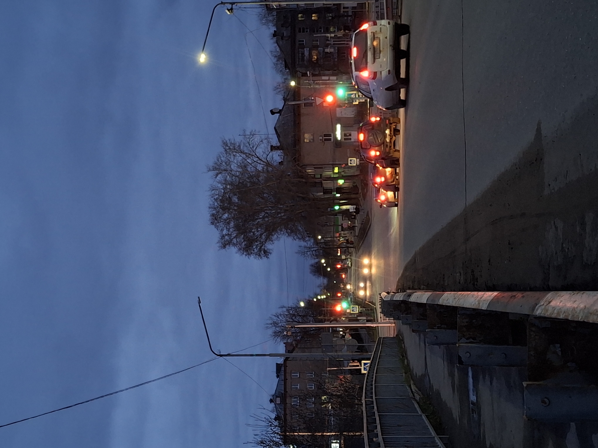 The image captures an urban street scene at dusk or early night, featuring a road flanked by buildings and trees. The sky is a deep twilight blue. The main subject is a multi-lane asphalt road on the right, where traffic is stopped at a red light. A white car is prominent in the foreground lane, its red taillights glowing brightly, followed by other vehicles further down the road, also displaying red taillights. Some vehicles also have illuminated headlights.
On the left side of the image, multi-story apartment buildings line the street, with lights visible in some windows, indicating occupancy. A large, bare deciduous tree stands in the midground on the left, its branches stark against the sky, suggesting late autumn or winter. Multiple streetlights illuminate the scene with a warm, yellowish glow.
Along the right edge of the road, there's a sturdy concrete barrier with a metal railing, marked with red and white stripes on its top, possibly separating the road from a pedestrian area or a lower level. Below this barrier, a concrete pathway or stairs are visible.
Notable details include several traffic lights displaying red circles and green arrows, indicating an intersection with specific turning lanes. A square road sign with a white background and a black symbol (likely a directional or regulatory sign) is visible near the buildings. The road surface appears damp, reflecting the vehicle and streetlights. The scene depicts a typical evening commute or activity in an urban setting, likely in Chaikovsky, Russia, as suggested by the architectural style of the buildings. No people are explicitly visible, and there is no legible text.