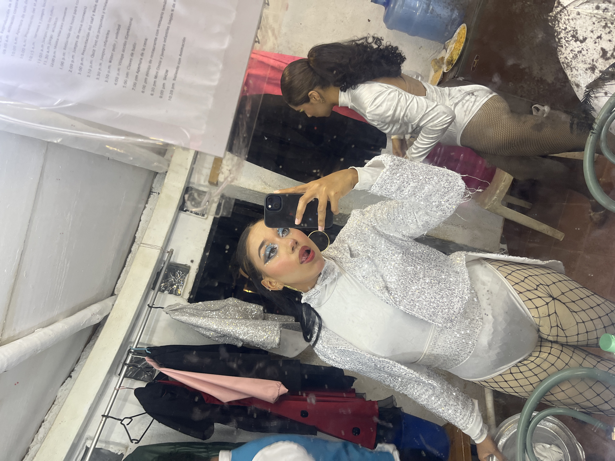 The image captures a mirror reflection of two young women within what appears to be a backstage area or dressing room in Porlamar, Venezuela. The primary subject, reflected in the foreground, is a woman with dark hair, detailed blue and white glitter eye makeup, pink blush, and red lipstick. She wears large gold hoop earrings and multiple ear piercings. Her attire consists of a white long-sleeved turtleneck bodysuit, a white sequined long-sleeved blazer, and black fishnet stockings. She is holding a black smartphone (likely an iPhone) to capture the image and has her tongue playfully extended. Her left hand is resting on a large, round, silver basin that contains fabric and a green hose or cable.
To the right, partially visible and facing away from the mirror, is a second woman with dark, curly hair tied in a high ponytail. She is wearing a white long-sleeved, high-necked bodysuit with an open back, and black fishnet stockings.
The background on the left features a metal clothes rack with several garments hanging, including a black jacket, a pink top, and a silver sequined item. Part of a blue and white garment is visible on the far left. The ceiling is white with exposed pipes. On the right, a dark surface, possibly a refrigerator or cabinet, holds a large blue water dispenser bottle, a smaller pink bottle, and a white plate containing yellow rice and fried items. A white plastic chair is positioned near the second woman. The floor is concrete or tiled, exhibiting some discoloration and debris. In the upper right section of the mirror's reflection, a laminated document listing a schedule of activities in Spanish is visible, with entries such as "SHOW" and "Clases de baile."