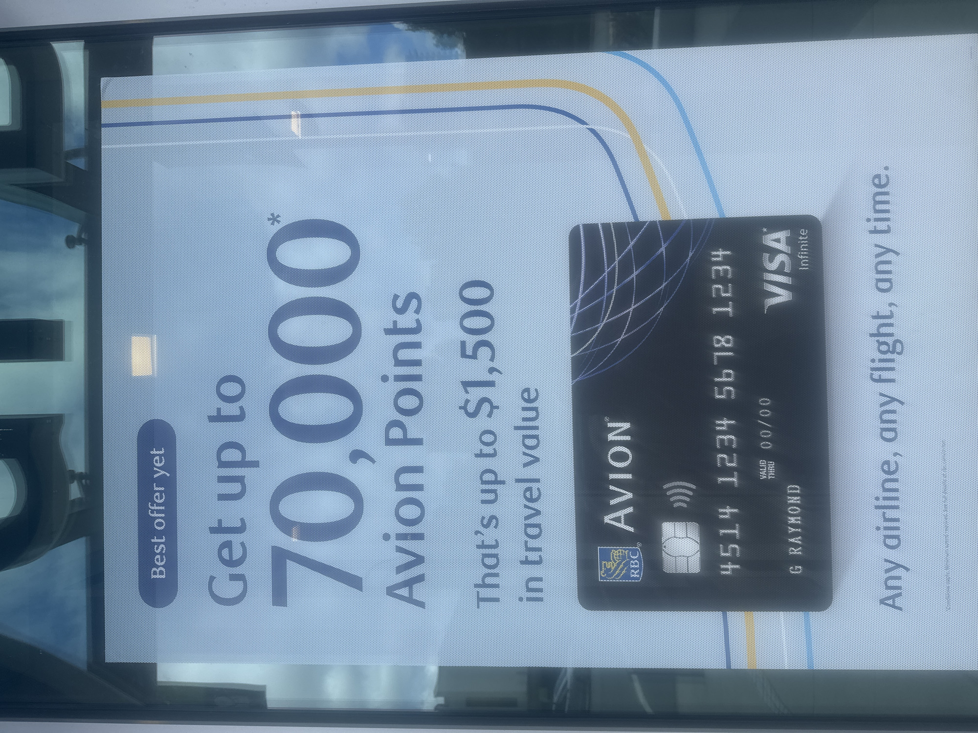 The image shows a close-up of a promotional poster displayed on a window, likely at a retail location or transit hub in Burnaby, Canada. The poster advertises an RBC Avion credit card with the headline "Get up to 70,000 Avion Points" and "That's up to $1,500 in travel value." Below this, a simulated RBC Avion Visa Infinite credit card is prominently featured with the name "G RAYMOND" and the last four digits of a card number "1234". The bottom of the poster reads "Any airline, any flight, any time." The background of the poster is a pale blue with abstract lines in blue and yellow. Reflections on the glass indicate an outdoor environment with a sky and possibly some buildings. The poster is adhered to a glass surface, suggesting it is a window display.