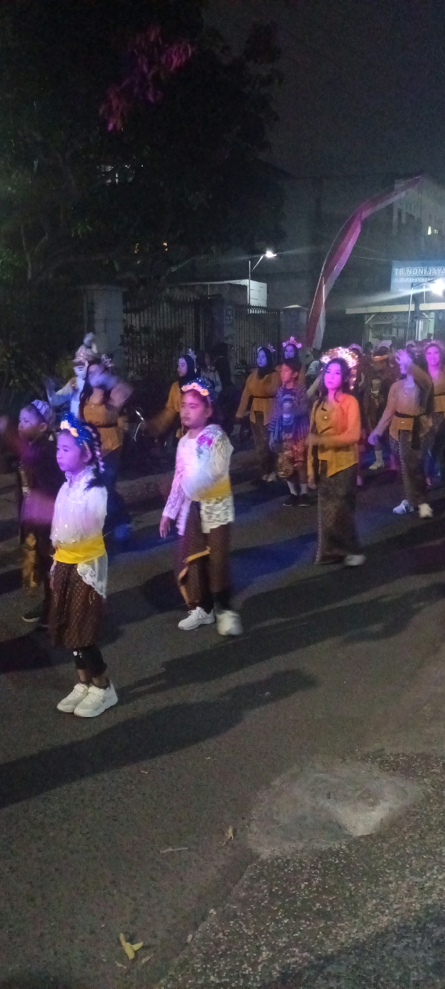 A group of people, primarily young girls, are walking in what appears to be a parade or procession at night. They are dressed in traditional or ceremonial clothing, with some wearing elaborate headpieces that are illuminated. The lighting is low, with artificial pink and blue lights casting a glow on the participants and the surroundings. In the background, there are buildings and trees, suggesting an urban or semi-urban environment. The ground is paved, indicating a street or road. The overall impression is one of a festive or cultural event taking place after dark.