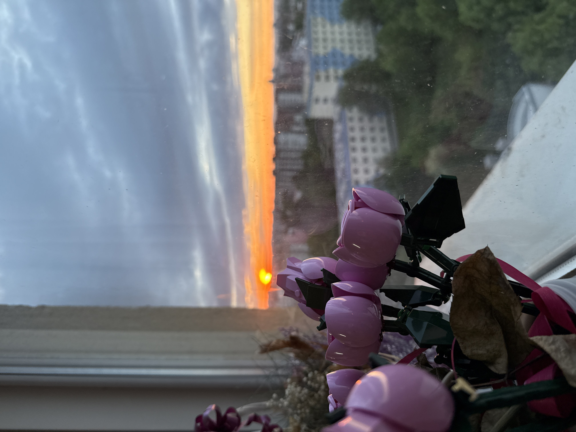 The image depicts a sunset viewed through a window, with pink artificial flowers in the foreground. The sunset is bright orange, contrasting with the cloudy gray sky on the left side of the frame. The sky has an orange glow reflected on the clouds. The buildings can be seen through the window on the right side, and are blurry. The pink flowers are clustered in the lower right corner, with plastic green stems. Dried flowers are arranged around the plastic flowers. A pink ribbon is woven into the arrangement. It is not possible to determine the specific location in Sofia, Bulgaria from the image.