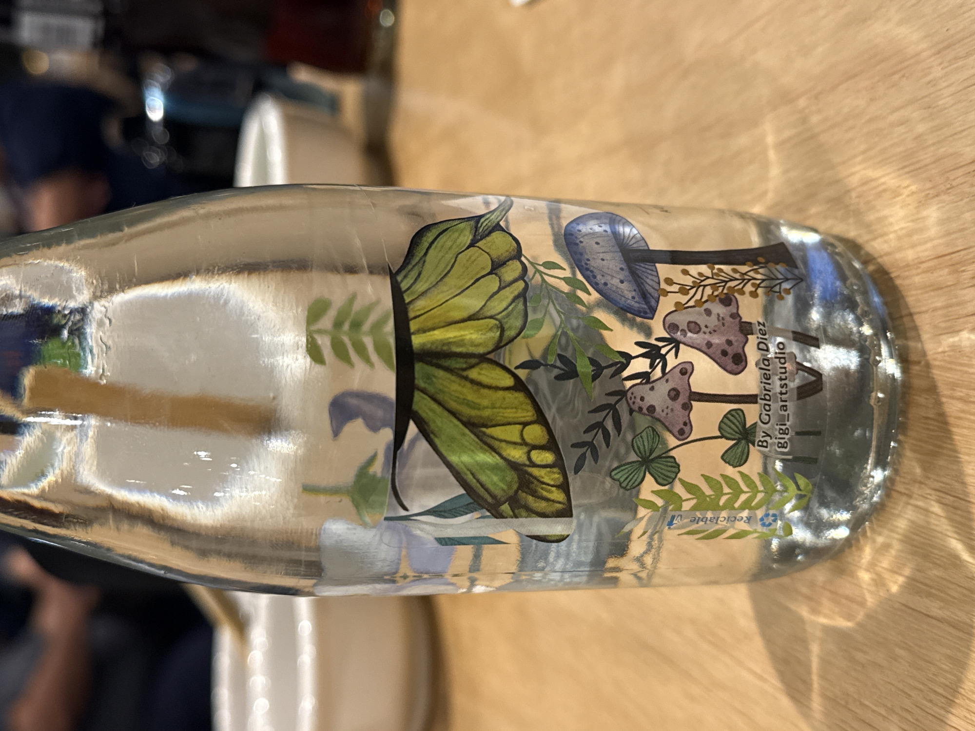 The image depicts a clear glass bottle decorated with a botanical illustration of mushrooms and a butterfly. The illustration also includes the text "By Gabriela Diez Gigi_artstudio" and a recycle symbol. The bottle is resting on a light-colored wooden surface. The photograph was taken indoors. The location, Querétaro, Mexico, is not visually confirmed in the image.