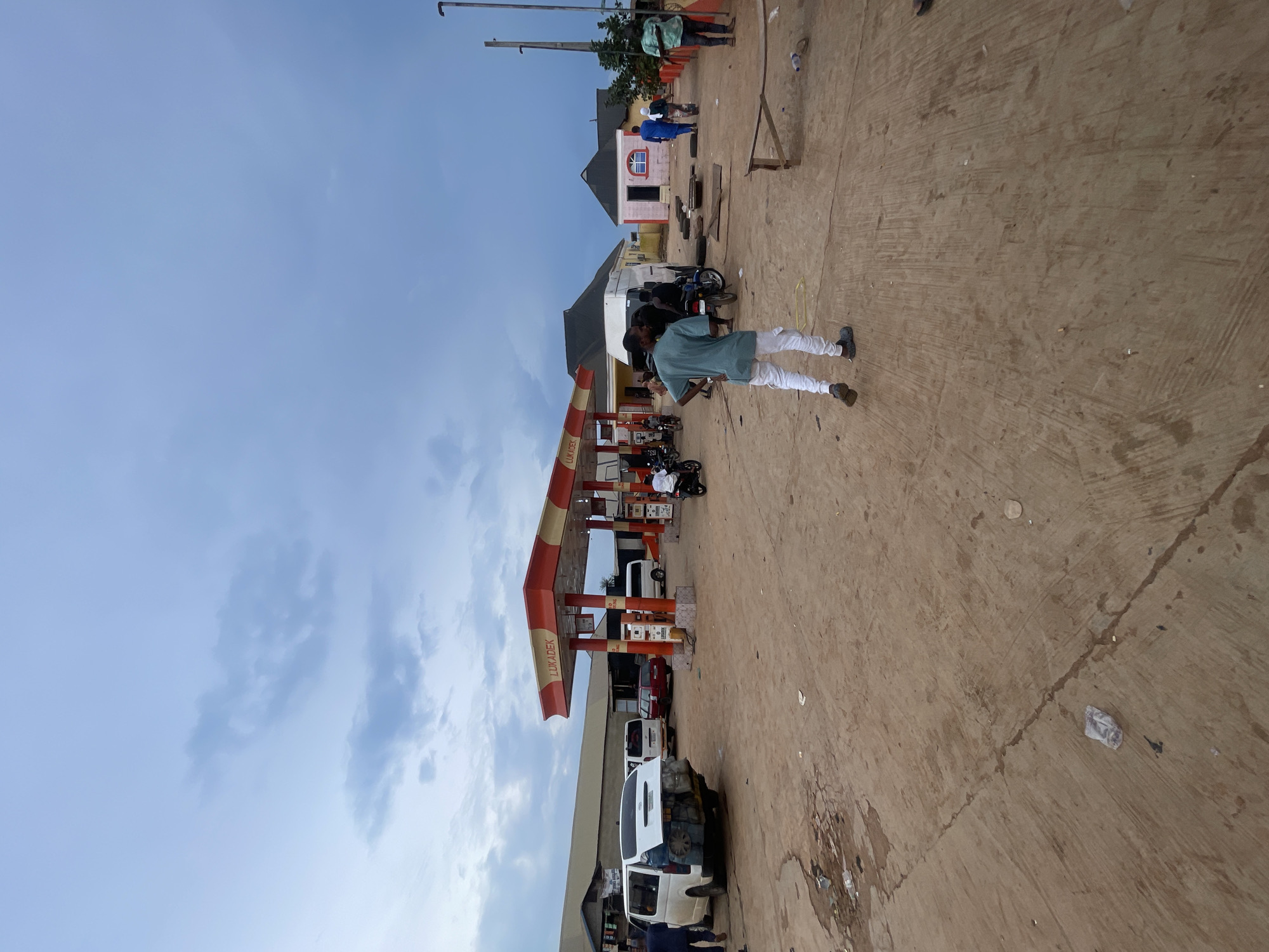 The image depicts a gas station in Osu, Nigeria. In the foreground, a person wearing a green shirt and white pants walks away from the camera. The ground is dusty and uneven, with some debris scattered around.
The gas station features a red and white awning with the name "LUKADEK" visible. Several vehicles are present, including a white van with luggage strapped to its back, a red car, and various motorcycles. Some people are standing around the gas station, while others are on motorcycles. Behind the gas station, there are buildings with corrugated metal roofs.
The sky is overcast with clouds, suggesting it may be evening or a cloudy day. The overall scene appears to be a busy, functional gas station in a rural or semi-urban area of Nigeria.