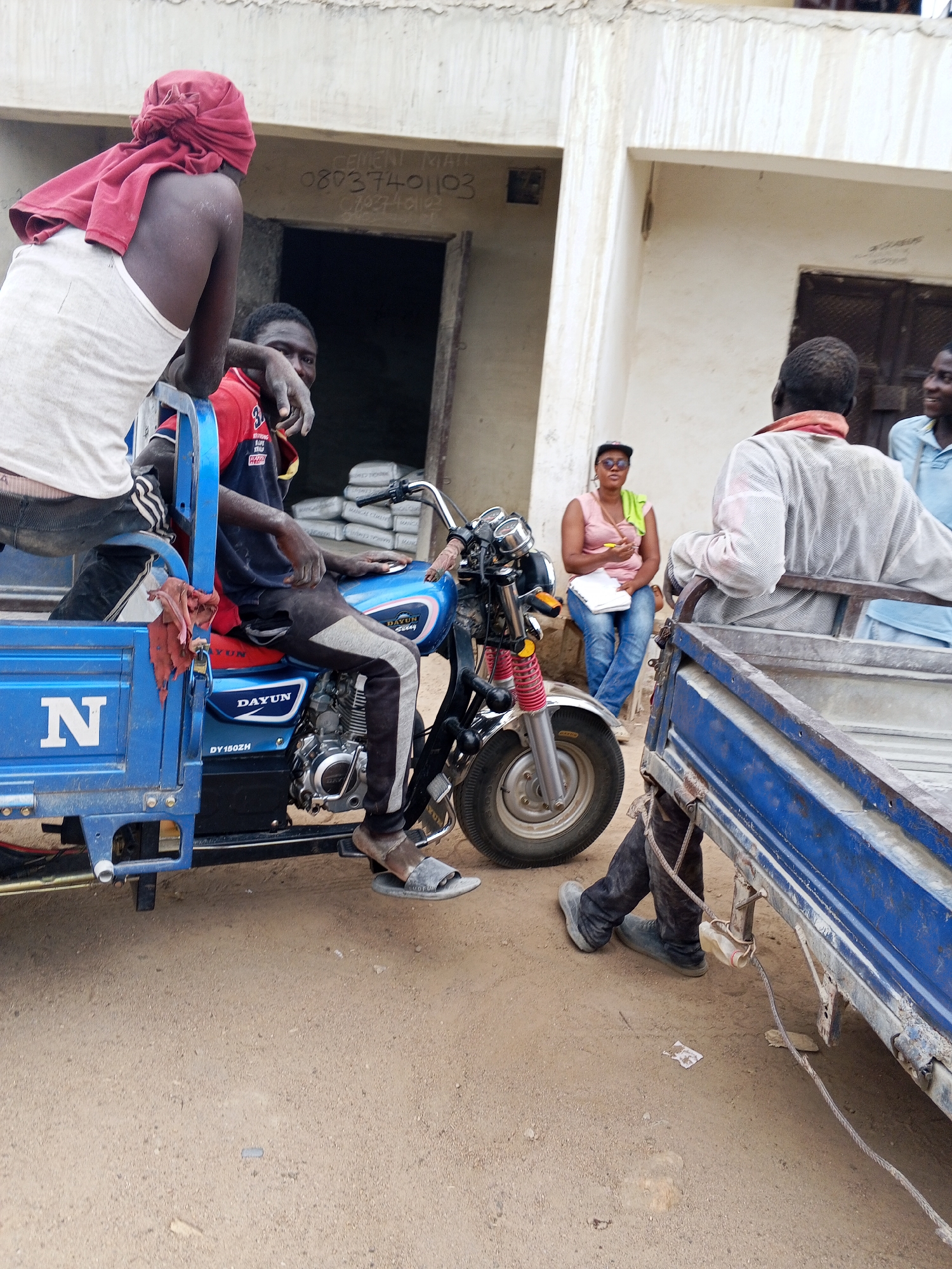 The scene is set in what appears to be a commercial area of Adankolo, Nigeria. Two men are positioned on a blue "Dayun" branded three-wheeled motorcycle, likely used for transporting goods. One man is seated on the motorcycle, while the other is seated on the sidecar. In the background, a woman is sitting on a stoop, appearing to be writing or taking notes. Another man stands to the right of the frame, partially obscured. Bags of cement are visible through the doorway of a building behind the motorcycle. The environment consists of a dirt or unpaved ground, and the buildings appear to be constructed from concrete or similar materials. Writing on one of the buildings is visible, reading "08037401103."