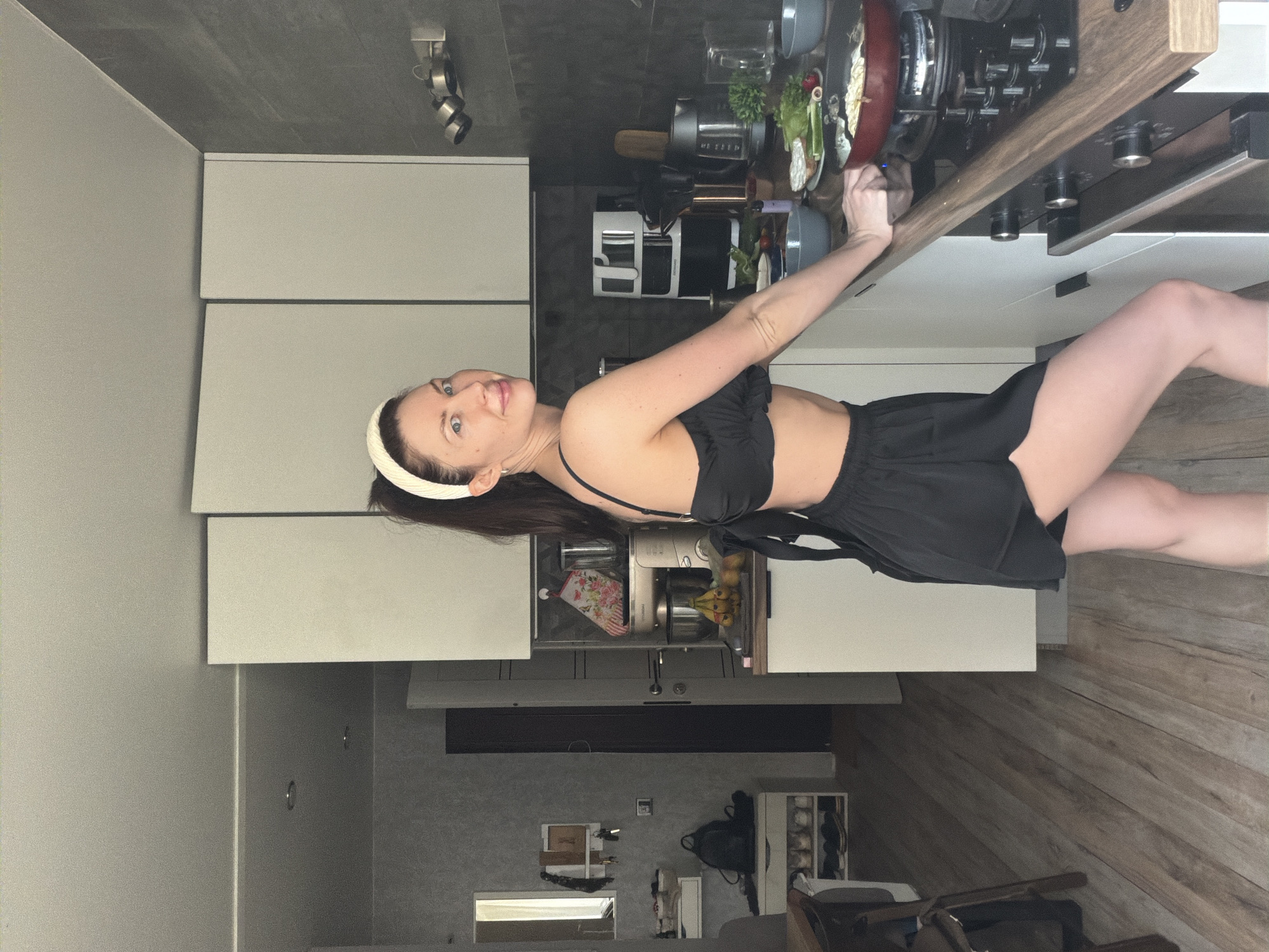 A woman is in a kitchen. She has brown hair and wears a white headband, a black top, and black shorts. She is standing to the right of a stove and counter, seemingly turning on the stove. There is a red pot on the stove with food in it. Other kitchen appliances are visible on the counter and in cabinets. The floor has a wood-like pattern. The kitchen appears to be in a home in the city of Perm, Russia.