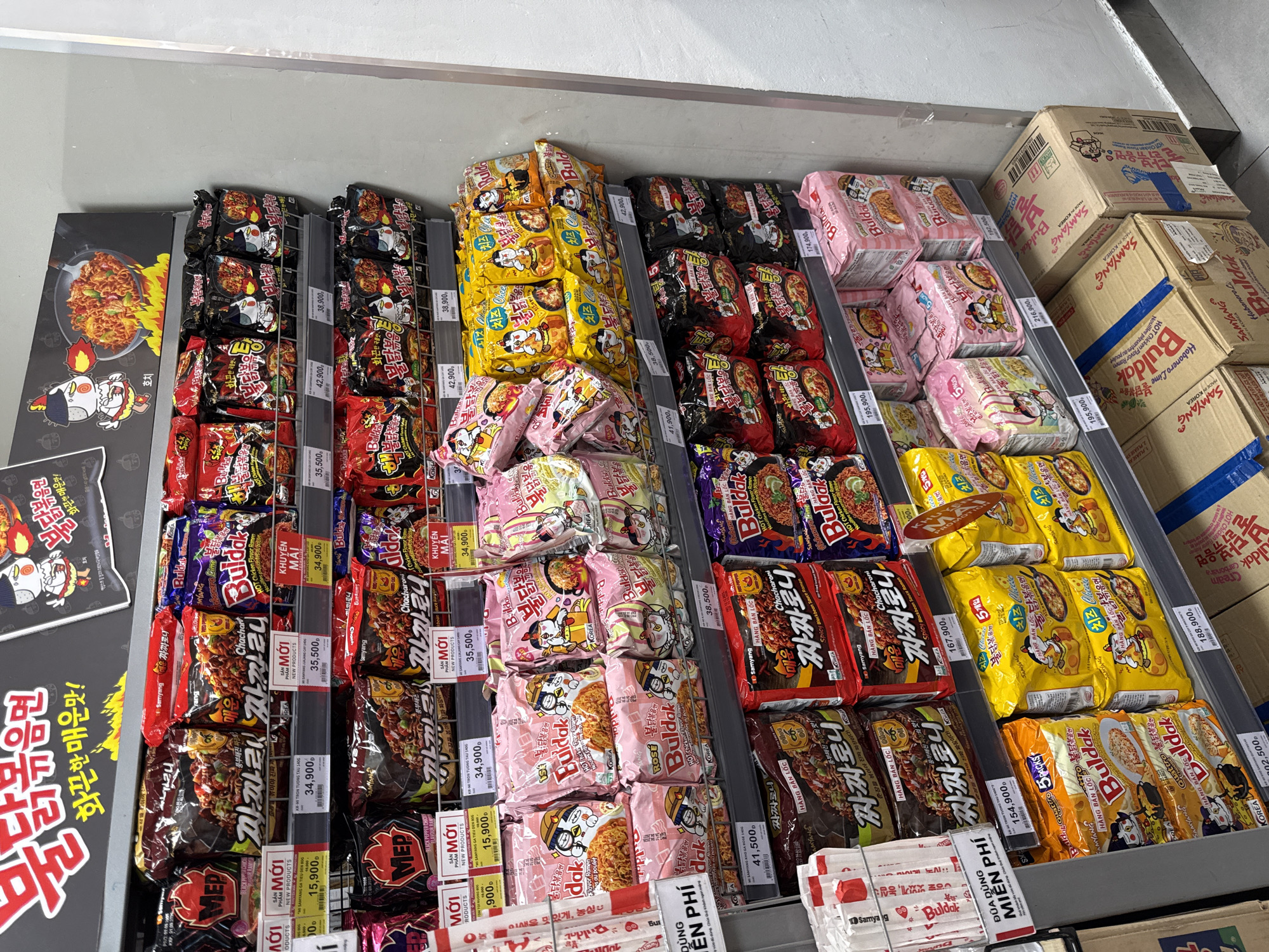 The image shows a shelf display of instant ramen noodles in a retail store. The products are predominantly Korean brands, with prominent packaging featuring cartoon characters and Korean text, alongside some Vietnamese text indicating promotions and prices. The shelves are organized with rows of noodle packets and larger multi-pack boxes. The background shows a plain, light-colored wall. There are no people visible in the image, and no actions or interactions are taking place. The environment appears to be a well-lit supermarket aisle. Based on the pricing and promotional language in Vietnamese, the location is likely a supermarket in Vietnam. It is impossible to pinpoint the specific city of Nha Trang without further contextual information.