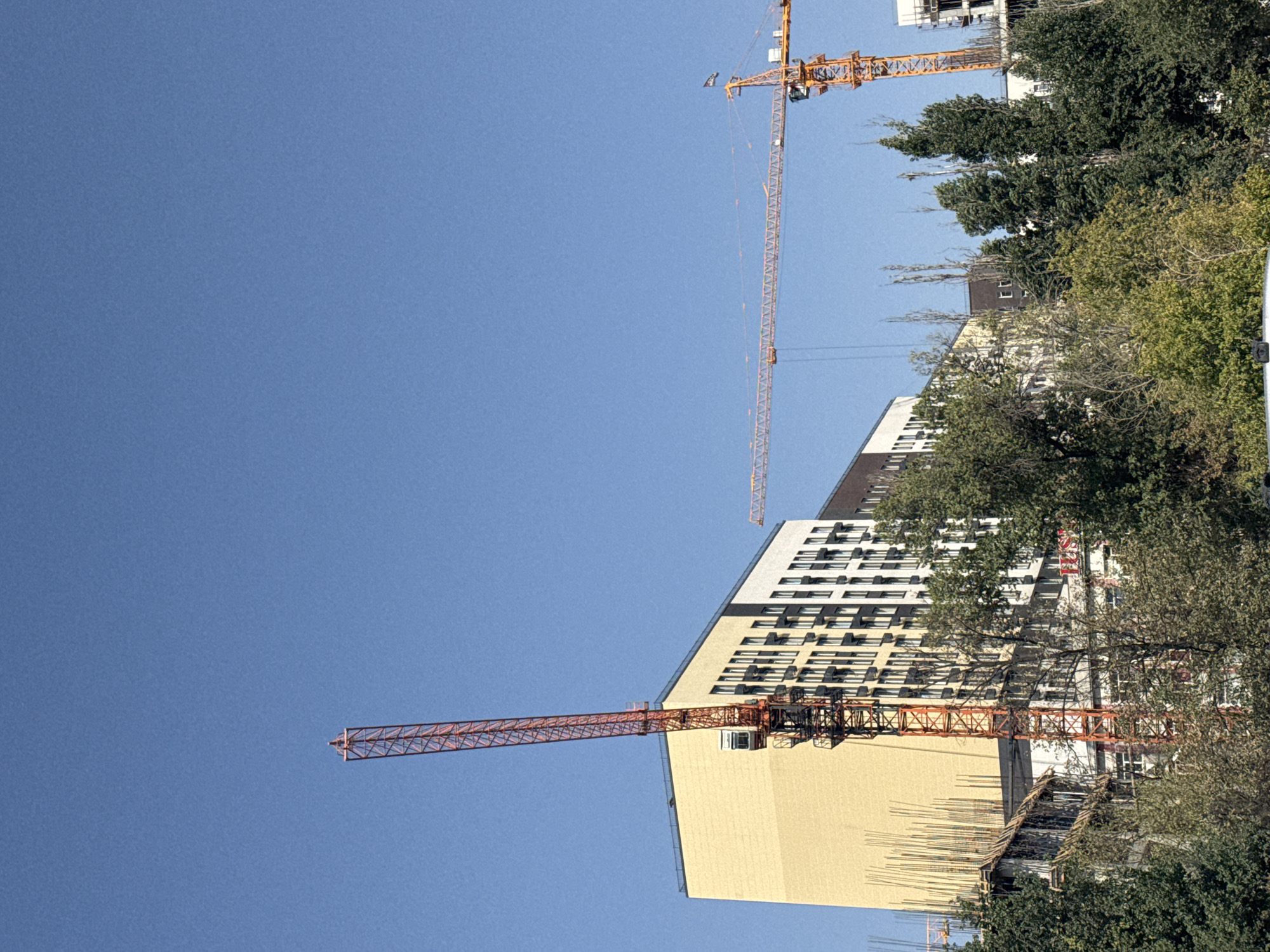 The image shows a construction site in Bishkek City, Kyrgyzstan, under a clear blue sky. Two construction cranes are visible. The crane on the left is red and prominently features a cabin and a tall vertical tower. To its right, a yellow crane with a long horizontal boom extends across the frame. In the background, a multi-story building with a light yellow facade and a section with white and dark windows is under construction. Trees, some with green foliage and others appearing more bare, are in the foreground, partially obscuring the lower portions of the buildings and construction site. The overall scene depicts an active urban development area.