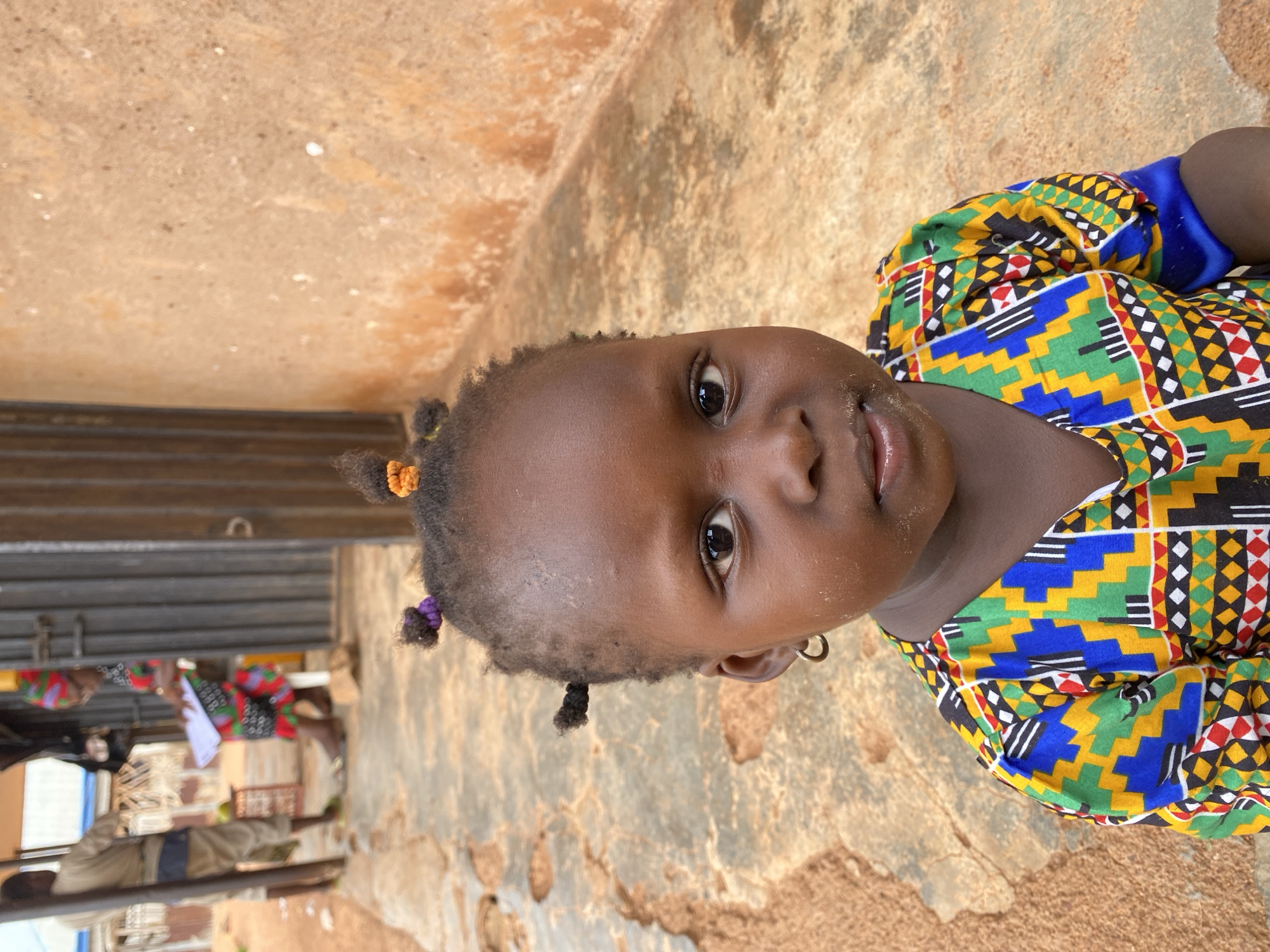 The image, taken in Kuru Dungus, Nigeria, features a young child, likely a girl, captured from the chest up, looking directly into the camera. She has dark skin and her hair is styled in small pigtails adorned with colorful bands, one orange and one purple. A small silver hoop earring is visible in her right ear. She wears a vibrant, short-sleeved top with a distinctive geometric pattern in shades of blue, green, yellow, black, and red, with white accents. Her face shows a slight dusting of dirt or sand, particularly around her mouth and forehead, and her dark eyes are wide and attentive, conveying a neutral yet engaging expression.
The setting appears to be outdoors, with an earthy, reddish-brown ground or wall forming the background, which is cracked and uneven in places. In the blurred background to the left, several other people are faintly visible, some also in patterned clothing, alongside what seems to be a metal gate or corrugated shutter and indistinct structures, suggesting a lively, possibly communal area such as a market or village street. The lighting is bright and even, indicating it is daytime. No discernible text is visible in the image.