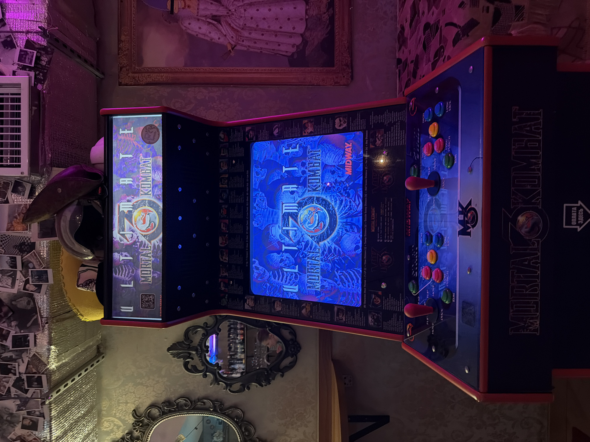 This indoor shot, taken in Moscow, Russia, features an "Ultimate Mortal Kombat" arcade cabinet as its central subject. The cabinet is powered on, displaying the game's title screen with the "MIDWAY" logo glowing in blue light. The top marquee also reads "ULTIMATE MORTAL KOMBAT."
The control panel of the arcade machine is visible, equipped with two red-handled joysticks and numerous colored buttons. Labels for the buttons include "HIGH PUNCH," "LOW PUNCH," "HIGH KICK," "LOW KICK," "BLOCK," and "RUN." Detailed instructions and character move lists are printed on the control panel, featuring text such as "All Characters Have Throws" and various distance indicators. The side of the cabinet is emblazoned with the "MORTAL KOMBAT" logo. A QR code is present on the front-facing side of the cabinet. A white label with the Russian text "ОПЛАТА ЗДЕСЬ" (meaning "PAY HERE") is affixed to the right side, indicating a payment point.
The arcade machine stands in a dimly lit room, illuminated by artificial light sources that cast a purple-pink hue across the scene. The walls are decorated with patterned wallpaper and feature ornate framed mirrors reflecting parts of the room. A large framed painting, possibly depicting a historical figure in period dress, hangs above the arcade machine. To the left, a collection of small black-and-white photographs is pinned to the wall next to a white air vent or cooler. A hat or cap is seen hanging below the vent. No people are visible, but the setup suggests an entertainment or gaming environment. The lighting suggests it is evening or night.