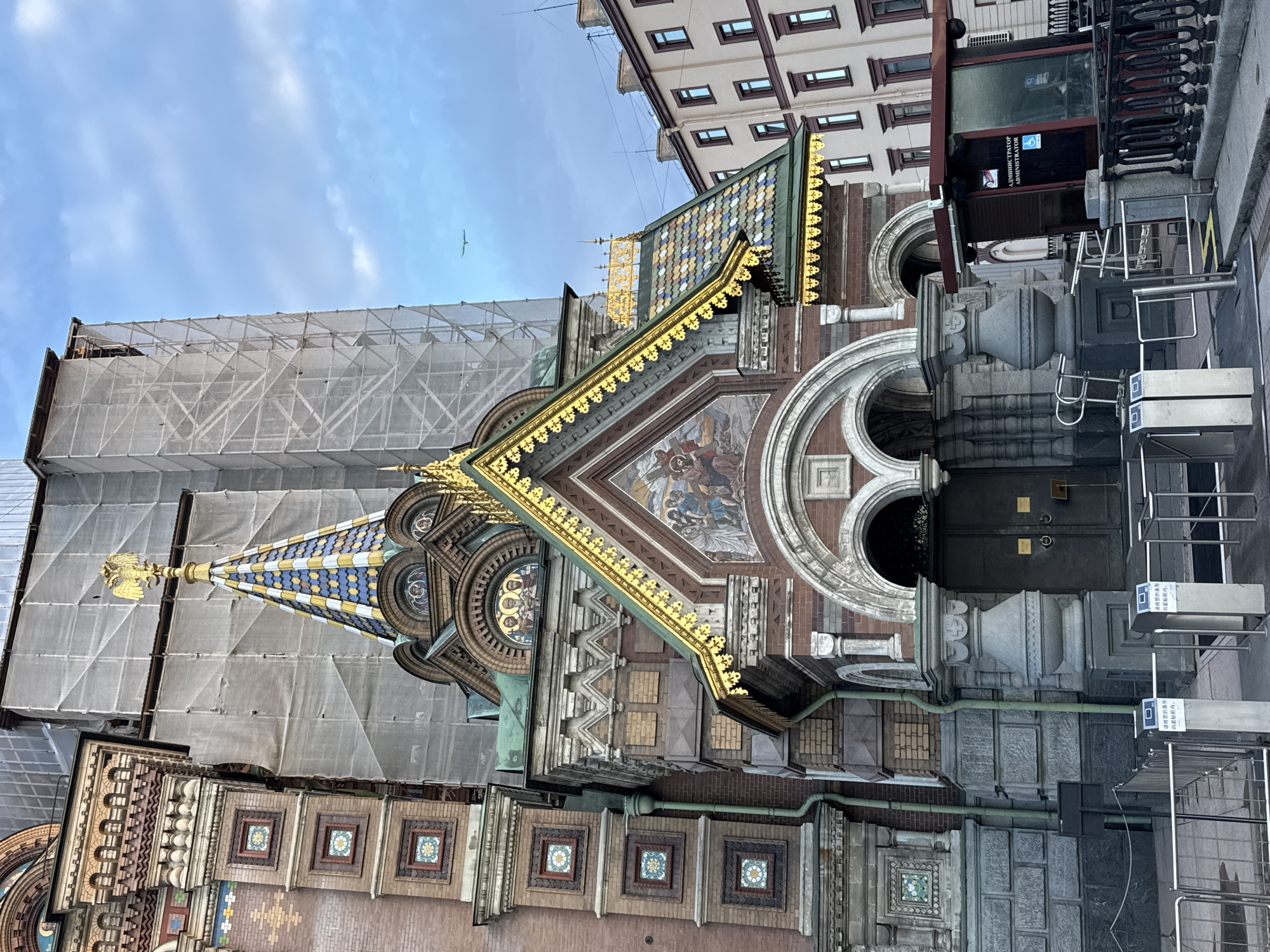 The image shows the Church of the Savior on Spilled Blood in Saint Petersburg, Russia. The church, a prominent landmark known for its ornate architecture, is partially covered by scaffolding and white tarpaulin, suggesting ongoing renovation or preservation work. The foreground features a metal turnstile system, indicating an entrance or controlled access point. A small kiosk with signage, including the word "ADMINISTRATOR" in English and Russian, is visible to the right. The sky is a pale blue with scattered clouds. The building's distinctive onion domes, intricate mosaics, and colorful tiled roof are partially visible, despite the scaffolding. The overall scene captures a blend of historical architecture with contemporary maintenance activities.