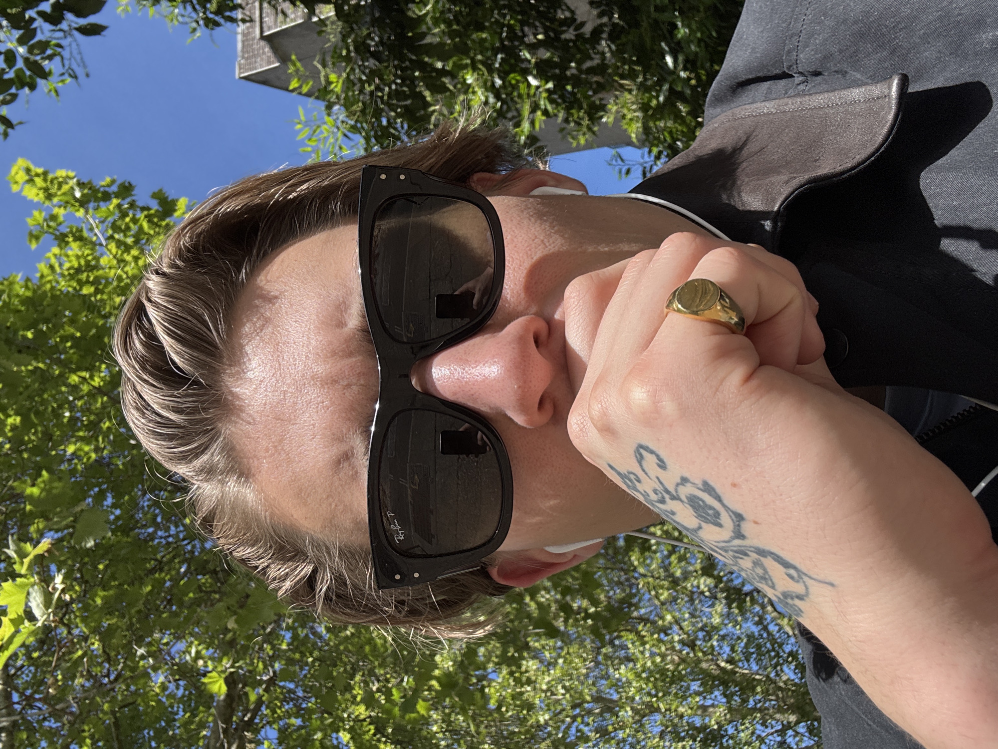A low-angle shot depicts a person outdoors on a sunny day. The individual wears dark sunglasses with the brand "Ray-Ban" visible on one lens. They also have white earbuds in their ears. The person's right hand is raised near their face, with a gold signet ring visible on their finger and a blue-ink floral tattoo on their wrist/forearm. The person wears a dark collared shirt or jacket. The background consists of green foliage (trees) and glimpses of a blue sky and buildings. The location appears to be London, United Kingdom, due to the architectural style of the buildings in the background and the overall aesthetic.