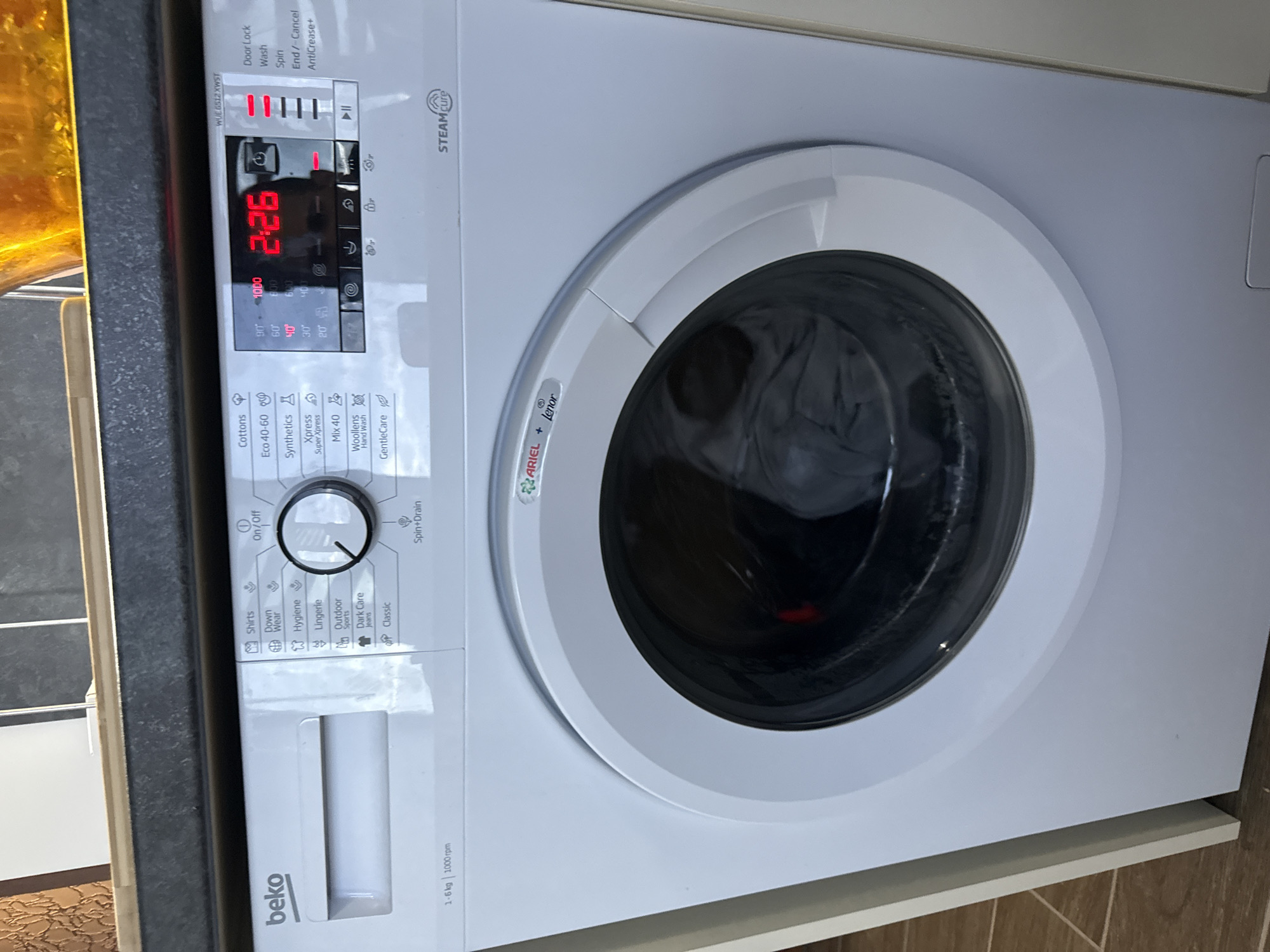 The image shows a white Beko washing machine with a digital display indicating a time of 2:26. The control knob is set to "On/Off," and other wash cycle options include Cottons, Eco 40-60, Synthetics, Xpress Super Xpress, Mix 40, Woollens Hand Wash, GentleCare, Shirts, Down Wear, Hygiene, Lingerie, Outdoor Sports, Dark Care Jeans, and Classic. The labels "ARIEL + Lenor" and "STEAM cure" are visible on the machine. The machine appears to be installed in a kitchen or laundry room setting with a dark countertop and wooden trim. The location in the city of Lovech, Bulgaria, is not ascertainable from the image.