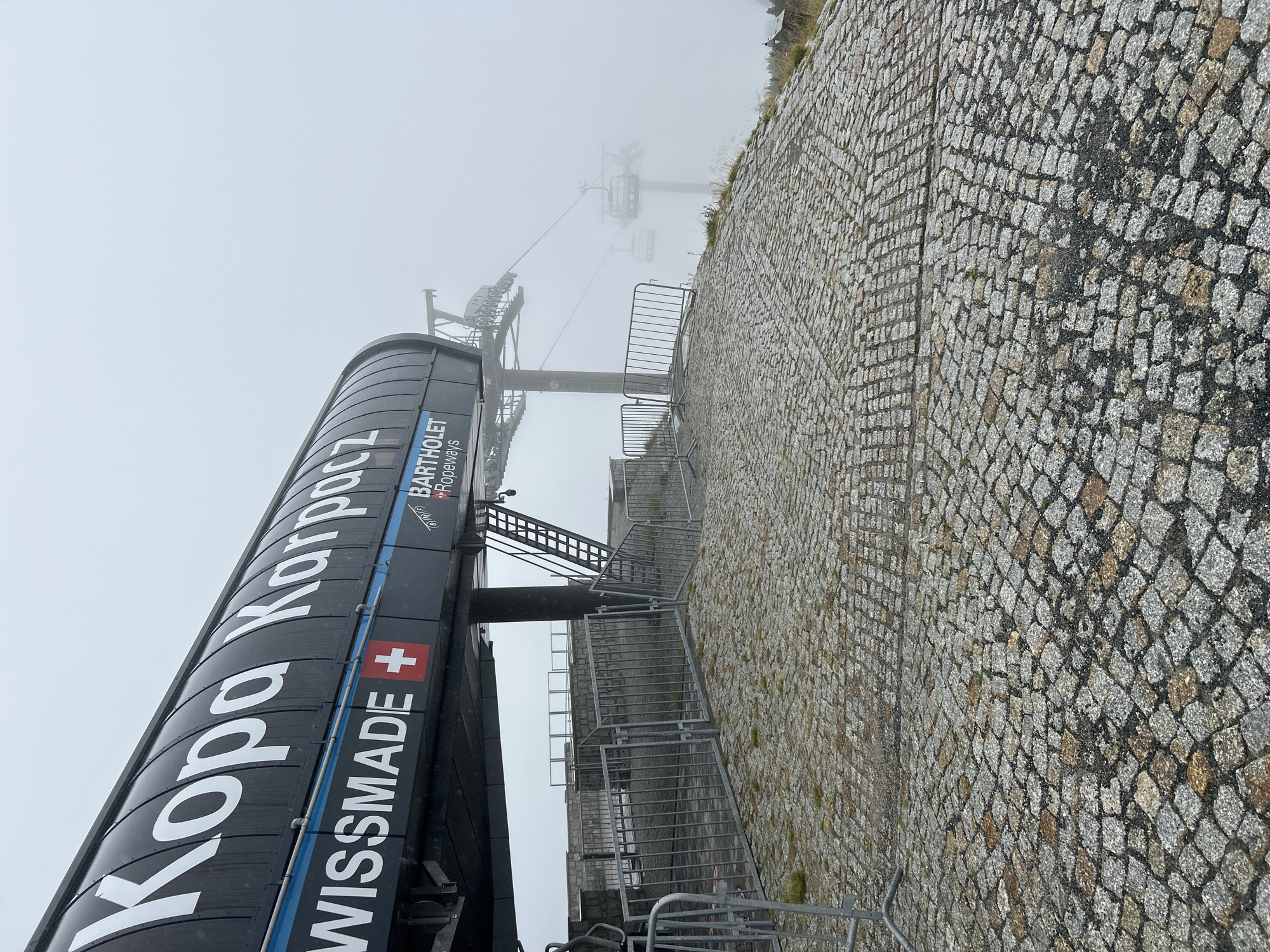The image shows the Kopa Karpacz ski lift station in Karpacz, Poland. The station is a modern structure with a black, curved roof displaying white lettering that reads "KOPA KARPACZ". Below this, the words "WISS MADE" are visible, followed by a Swiss flag symbol. The ski lift mechanism, including cables and chairs, is visible disappearing into the fog. The foreground is covered in cobblestone paving. Metal barriers are set up around the station, suggesting a controlled access area. The overall atmosphere is overcast and foggy, obscuring the background landscape.