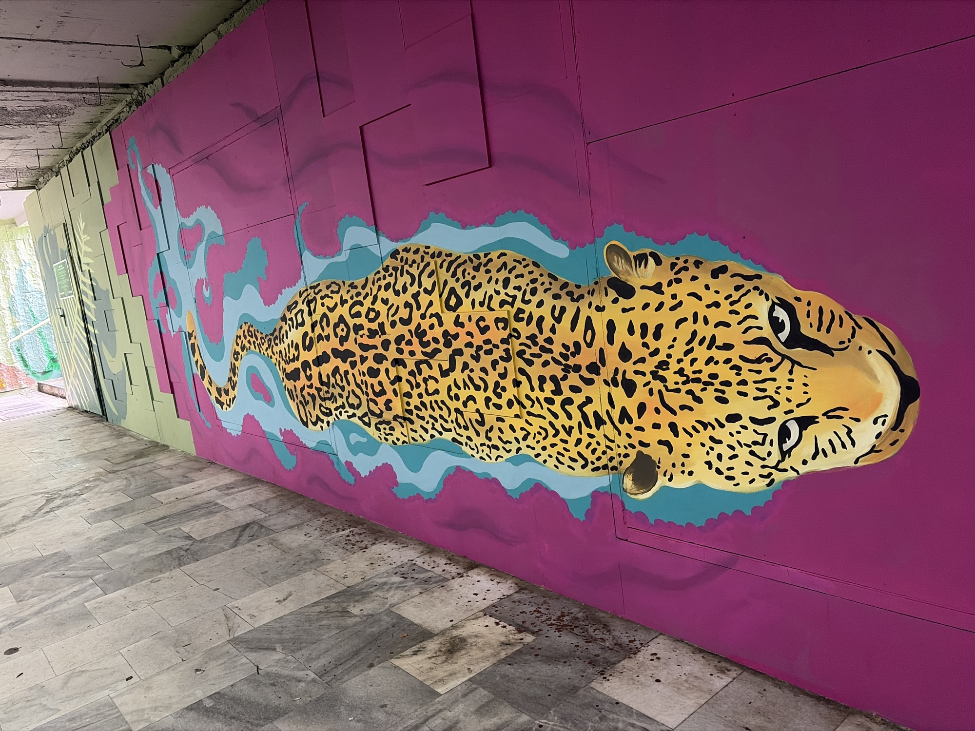 The image depicts a mural of a jaguar on a wall in an urban environment, likely in Sofia, Bulgaria. The mural features a stylized jaguar with yellow and black spots outlined in turquoise. The wall is painted a bright purple and the mural is on a wall composed of panels. To the left of the jaguar, the mural transitions to a green section. The floor is tiled with grey marble tiles, showing signs of staining or dirt. The tunnel has overhead concrete beams. The scene appears to be indoors, possibly a pedestrian underpass or walkway. The lighting is subdued. There are no people visible in the frame.