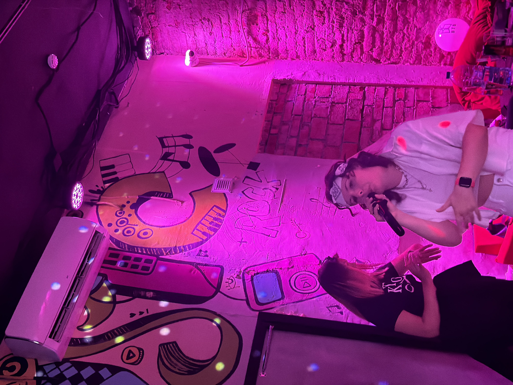 The image depicts an indoor scene illuminated by pink and purple lights, suggesting a nightlife or entertainment venue. A person in the foreground is holding a microphone and appears to be singing or performing. They are wearing a white open shirt over a white t-shirt, white pants, and a white bandana around their head, along with a smartwatch on their left wrist. To the left of the performer, a second person in a black t-shirt and black skirt is standing and looking towards the performer. The background features a wall with colorful graffiti depicting musical notes, a cassette player, and abstract shapes. An air conditioning unit is mounted high on the wall. Additional pink lights are visible on the ceiling. A bottle of water and a pink balloon with a logo are on a table in the lower right portion of the image. The brick texture of the wall is visible in the background, indicating a potentially industrial or rustic interior. The overall atmosphere appears lively and celebratory. There is no information available to determine the specific location in Saint Petersburg, Russia, or any specific city for this image.