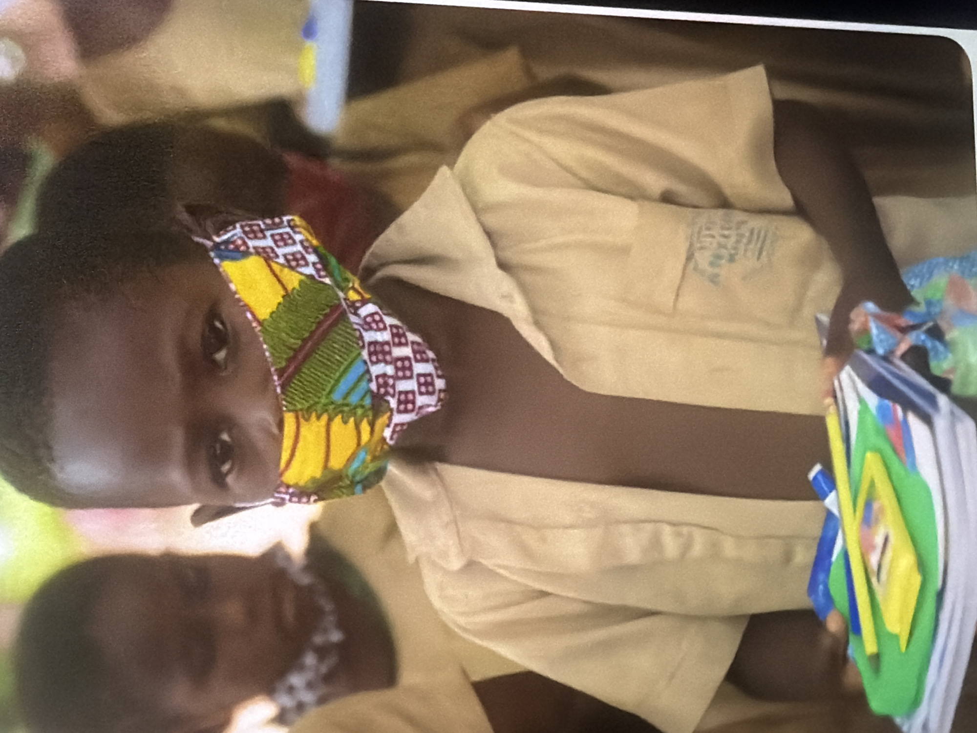 A young child wearing a tan school uniform and a colorful fabric face mask is the central subject of this close-up, medium shot photograph. The child's eyes are looking directly at the camera, and they are holding a stack of books and school supplies. The books are stacked on top of each other, with a green folder, a yellow pencil, and a blue marker visible on top. The child's right arm is extended, holding the stack of supplies. The background is blurred, but suggests other children in similar uniforms are present, indicating a school or educational setting. The lighting is natural and appears to be outdoors.