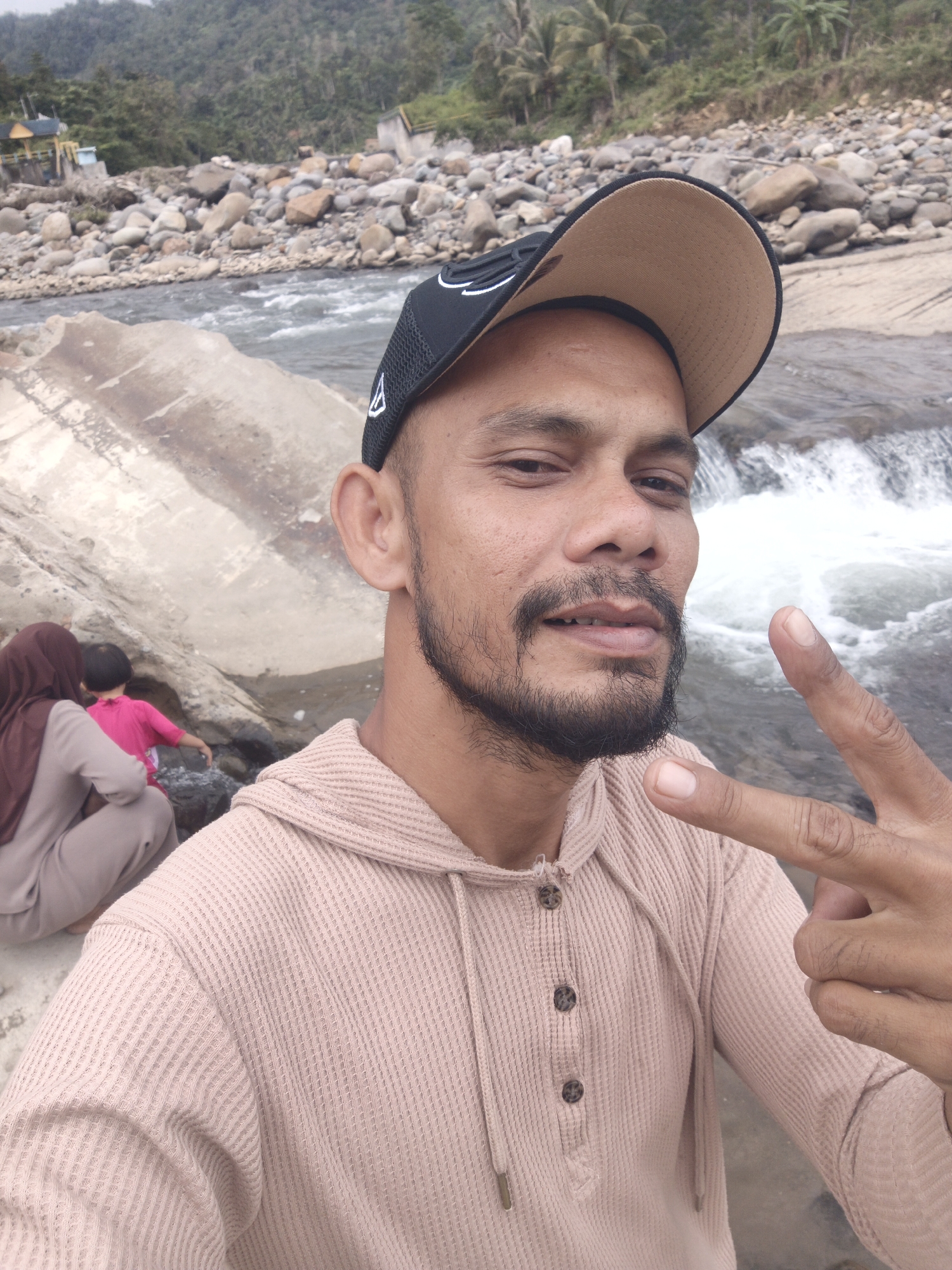 An adult male in Meurah, Indonesia, takes a selfie by a river. He has dark skin, a short beard, and wears a black baseball cap with a tan brim and a light pink waffle-knit hooded henley shirt. His right hand is raised, forming a "peace" sign. In the mid-ground, a woman wearing a reddish-brown hijab and a light brown outfit is seated near large rocks on the riverbank, facing away from the camera. Beside her, a child in a bright pink shirt is near the water. The background features a river with visible whitewater, a rocky shore, and dense green mountains or hills with some scattered palm trees and small structures. The sky appears overcast.
