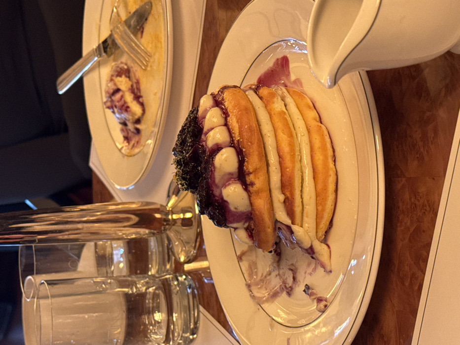 The scene is set at a dining table within an upscale restaurant in the city of Unknown, Unknown. In the foreground, a stack of four pancakes, layered with a light-colored cream or custard and topped with dark purple berry compote and a green garnish, rests on a white plate with a blue and gold double-line rim. Streaks of the purple compote are visible on the plate. To the lower right, a white ceramic creamer with a silver rim stands on the polished, reddish-brown wooden table. In the midground, two clear glass tumblers filled with water and a tall, golden, reflective decorative object are present. In the background, a second white plate with a similar rim displays remnants of a consumed meal, along with a silver fork and knife resting on it. A person wearing dark clothing is partially visible in the upper right background. No discernible actions or interactions are actively occurring, but the partially eaten meal suggests recent dining.