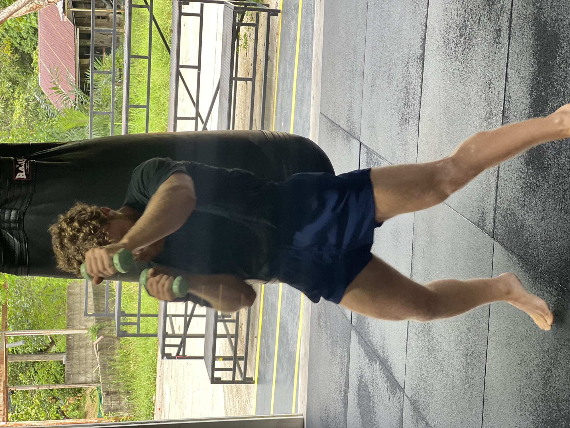 A man with curly brown hair is exercising with two green dumbbells in front of a large black punching bag. He is wearing a dark t-shirt and navy blue shorts. The man is in a gym with a dark gray tiled floor. In the background, there are metal benches and some vegetation, suggesting an outdoor or semi-outdoor gym. The scene appears to be in Chalong, Thailand, based on the context of other images. The man is in motion, suggesting he is actively performing a workout routine.