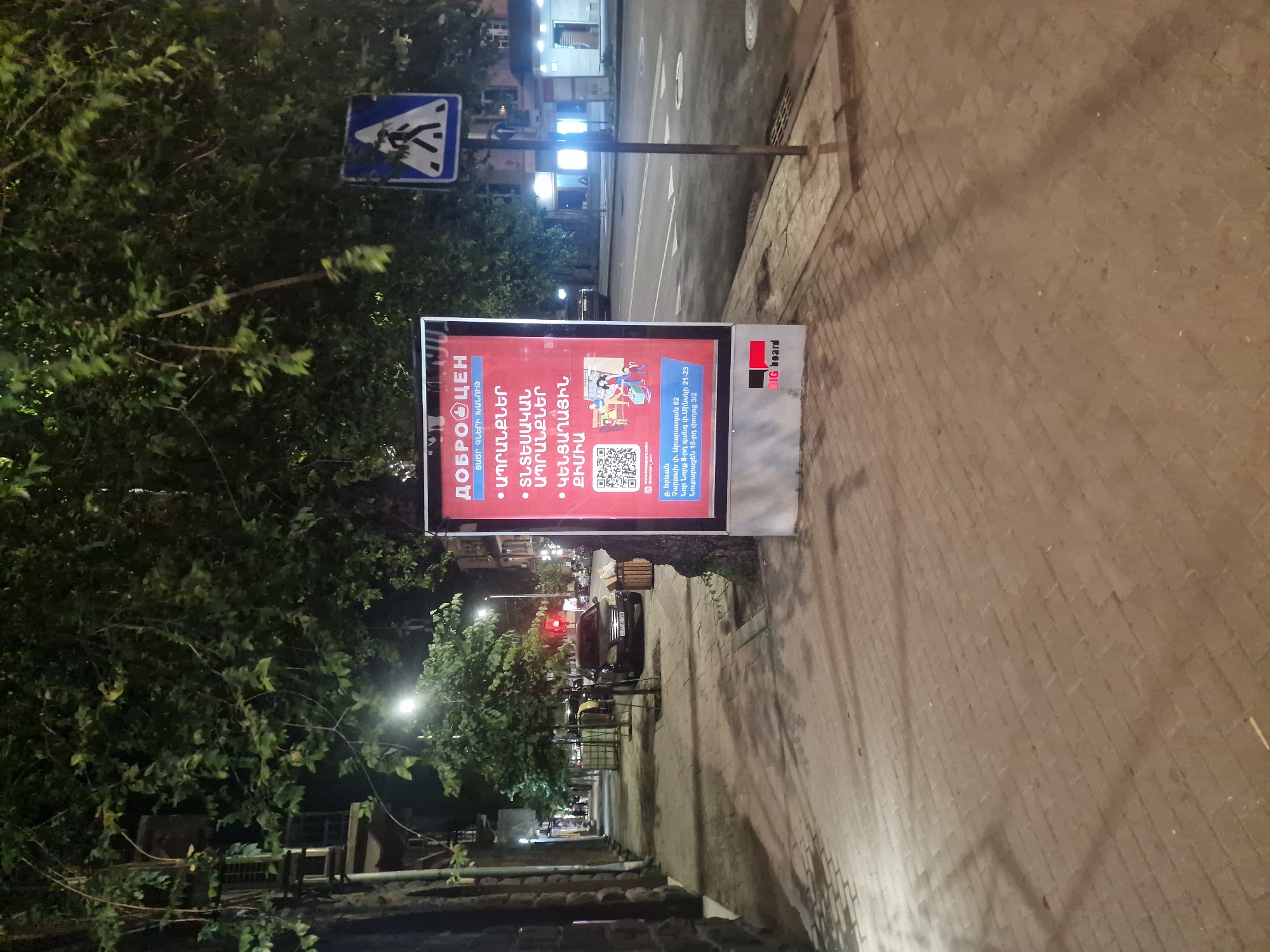 This media file shows a street scene at night, with a focus on a brightly lit advertisement kiosk on a sidewalk. The kiosk displays a red advertisement with text in both Cyrillic and Armenian. The Cyrillic text "ДОБРОЦЕН" is prominent at the top of the advertisement, likely a brand name. Below this, Armenian text lists categories such as "PRODUCTS," "HOUSEHOLD GOODS," and "HOUSEHOLD CHEMICALS." A QR code is also visible on the advertisement, along with what appears to be a web address or social media handle.
The setting appears to be an urban environment, with a street running alongside the sidewalk. Buildings with illuminated windows are visible in the background, suggesting businesses or residences. A pedestrian crossing sign is mounted on a pole near the advertisement. Trees with dense foliage partially obscure the view, creating shadows on the sidewalk. A dark-colored car is parked on the street, and streetlights cast a warm glow on the scene, indicating nighttime. The sidewalk is paved with bricks. The overall impression is that of a typical urban street at night, with commercial advertising playing a significant role.