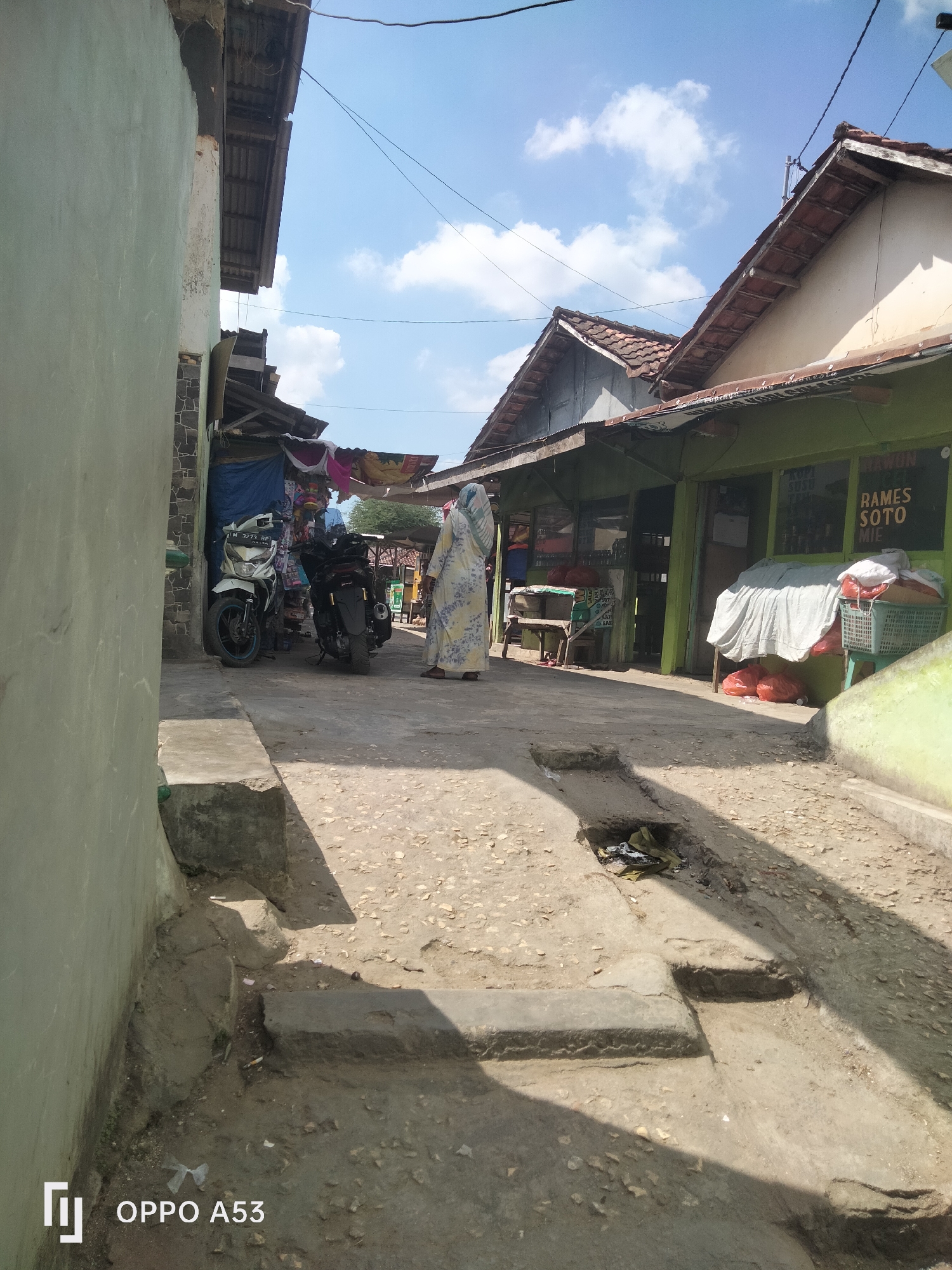 A street scene in what appears to be a rural area of Badung, Indonesia, is depicted. The image shows a narrow dirt road with a concrete drainage ditch running alongside it. On the right side of the road, there is a row of small shops or stalls with green facades and tiled roofs. One shop has a sign that reads "RAMES SOTO MIE." A white sheet covers some items on a table in front of this shop, and red plastic bags are visible on the ground. A person wearing a traditional patterned headscarf and a long, light-colored dress is standing near the shop. On the left side of the road, a light-colored motorcycle and a dark-colored motorcycle are parked. Further down the road, there are more stalls and buildings. The sky above is blue with scattered white clouds. The overall impression is of a modest commercial area in a developing region.