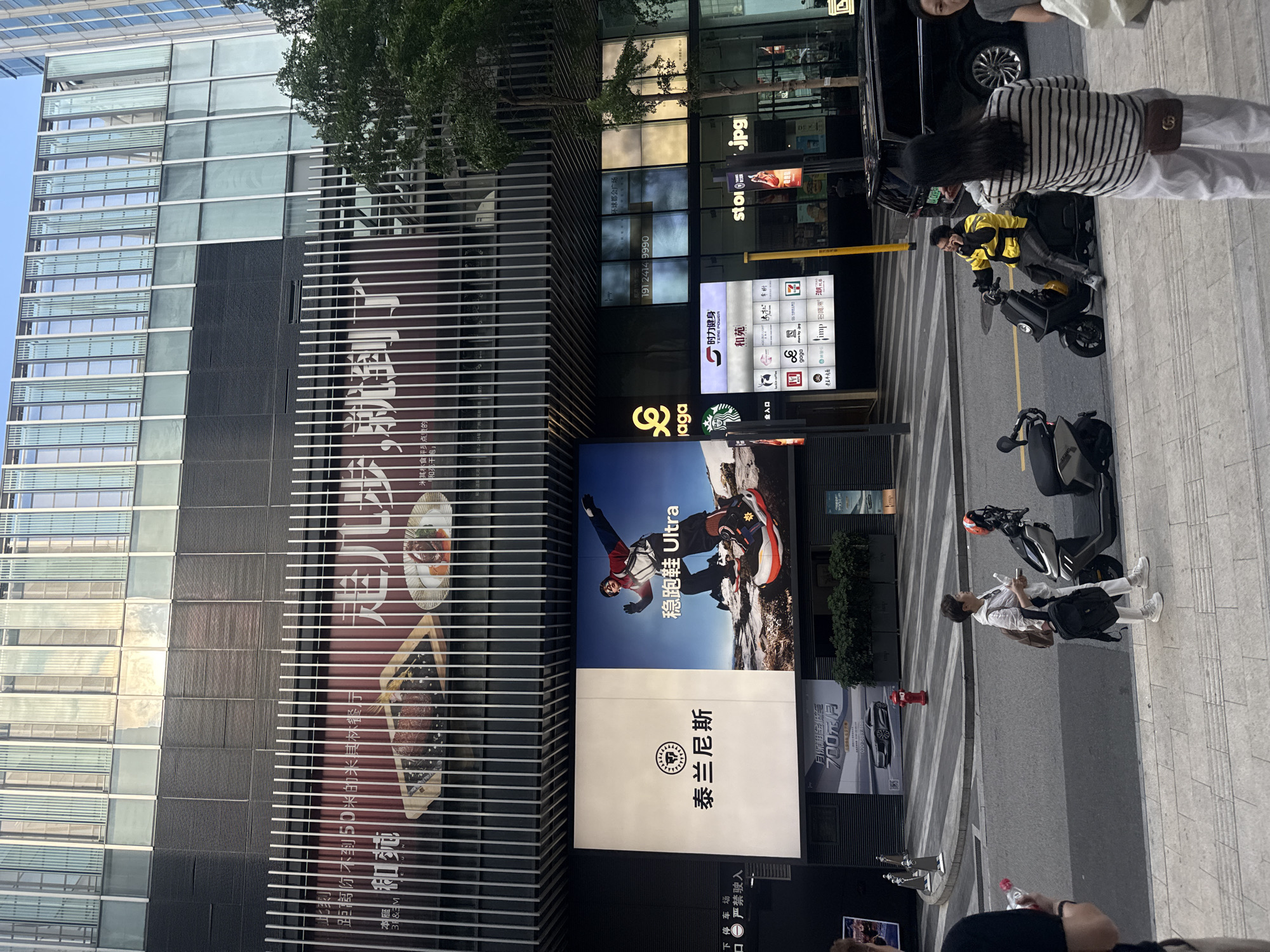 This media file presents a vibrant urban street scene in front of a modern high-rise commercial building, likely located in Haizhu District, China. The photograph was taken on a bright, sunny day, indicated by the clear blue sky and distinct shadows.
The scene features several individuals and typical city elements. On the sidewalk, a person in a black and white striped shirt and white pants is seen from behind, carrying a small brown shoulder bag. Further down, a person dressed in all white stands near a parked electric scooter. In the street, a delivery rider wearing a bright yellow jacket is seated on an electric scooter. A black car is partially visible, parked near the curb. Multiple electric scooters are parked or being ridden, a common sight in Chinese cities.
The building's facade is a mix of glass panels and advertising displays. A large, vertically oriented red banner on the left side of the building advertises a "Michelin restaurant" less than 50 meters away, with text reading "距离你不到50米的米其林餐厅" and "和苑" (He Yuan), which appears again on a smaller sign. Below this, a prominent digital billboard displays an advertisement for athletic footwear, featuring a person in hiking gear and the text "稳跑鞋 Ultra" (Stable running shoes Ultra) and "泰兰尼斯" (Terra Nova/Tairannis).
Various retail and service establishments are indicated by illuminated signs and logos, including Starbucks, 7-Eleven, KFC, Luckin Coffee, Dairy Queen (DQ), CoCo Tea & Juice, Pizza Hut, Baskin Robbins, McDonald's, Burger King, Adidas, Nike, '时力健身' (Power Fitness), and 'gaga'. A sign also points to '环球都会广场' (Global City Plaza) and displays a phone number "191 2414 9990". Another sign advertises "月保租金低至 700元/月" (monthly rental as low as 700 yuan/month), suggesting commercial leasing opportunities. A warning sign for an underground parking entrance reads "下停车场 严禁驶入" (Underground parking, no entry). The street itself has clear markings, including painted crosswalks and a yellow dividing line.