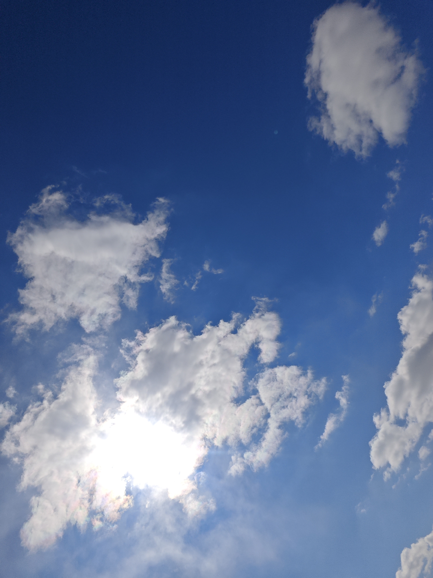 The image shows a view of the sky with scattered clouds. The sky is a bright blue color. There are a few fluffy white clouds spread throughout the frame, one of which is obscuring the sun, creating a bright glare. The clouds are mostly white, with some grey shading on their undersides. The location is unknown.
