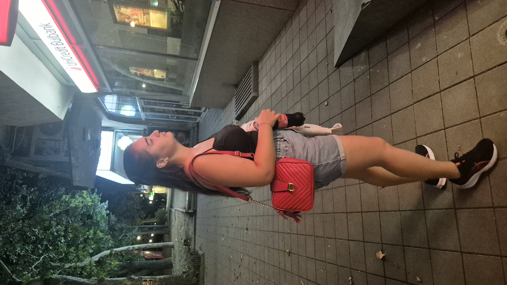 A woman stands on a paved sidewalk in front of a UniCredit Bulbank. She is wearing a black tank top, denim shorts, and black athletic shoes. A red crossbody bag with a gold clasp hangs on her right side, and she holds a dark-colored bottle in her right hand. The woman is looking towards the bank's windows. Trees line the sidewalk to her left, and a street lamp is visible in the background. The environment suggests an urban setting at dusk or night.