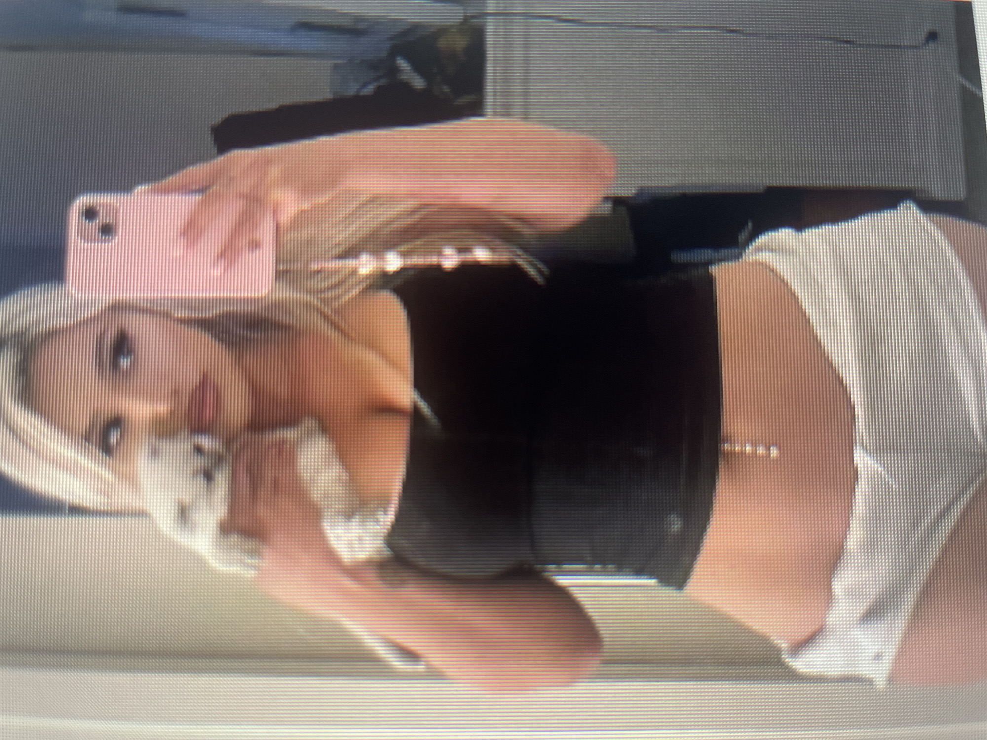 A blonde woman is taking a selfie in a bathroom. She is holding a small white dog in her left arm and her right hand is holding a pink iPhone. She is wearing a black tank top and white shorts. There is a pearl necklace hanging from the iPhone. She is wearing a belly chain. The background appears to be a bathroom with a tiled wall.