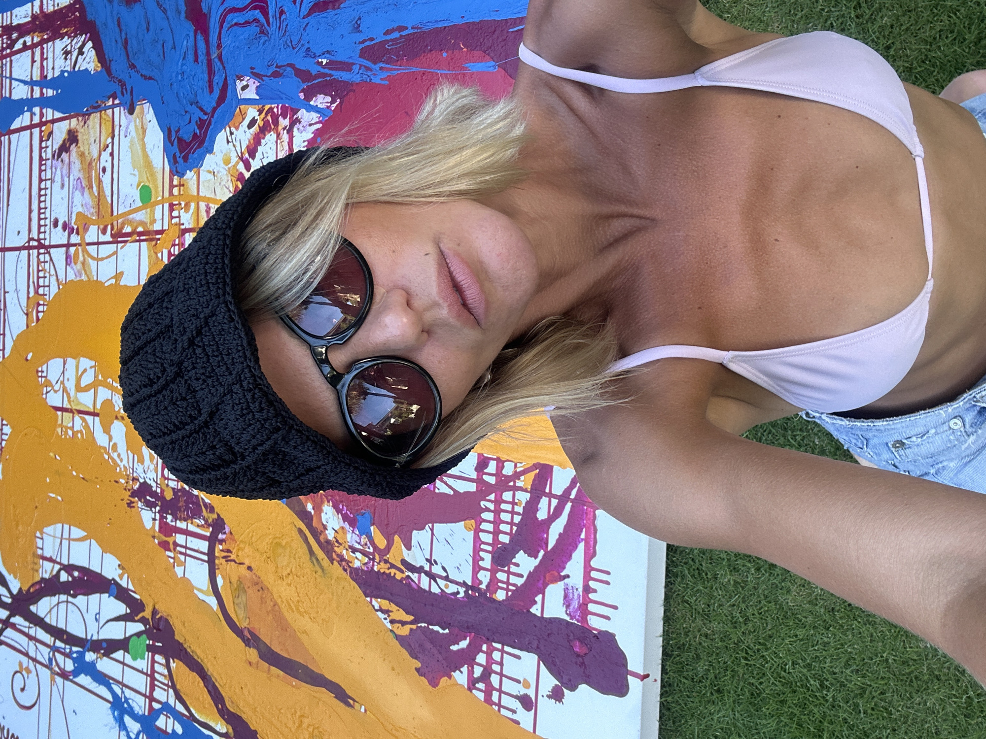A woman wearing a light pink bikini top, light blue denim shorts, a black crocheted beanie, and dark sunglasses is posed in front of a large abstract painting. The painting features a white background with splatters of blue, yellow, purple, pink, and orange paint. The woman is lying on a patch of green grass. The location is outdoors in the city of North Kuta, Indonesia.