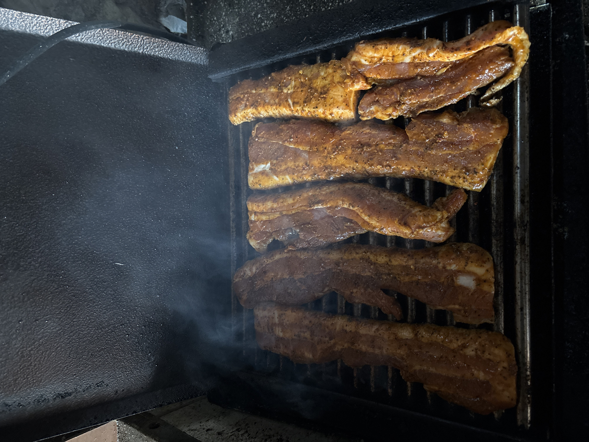 The image shows several pieces of meat being cooked on a grill inside what appears to be a smoker. The meat is arranged in a row on the grill grates. Smoke is visible, particularly on the left side. The interior of the smoker is black, with some parts showing texture. There is no evidence to support the location being Chirpan, Bulgaria.