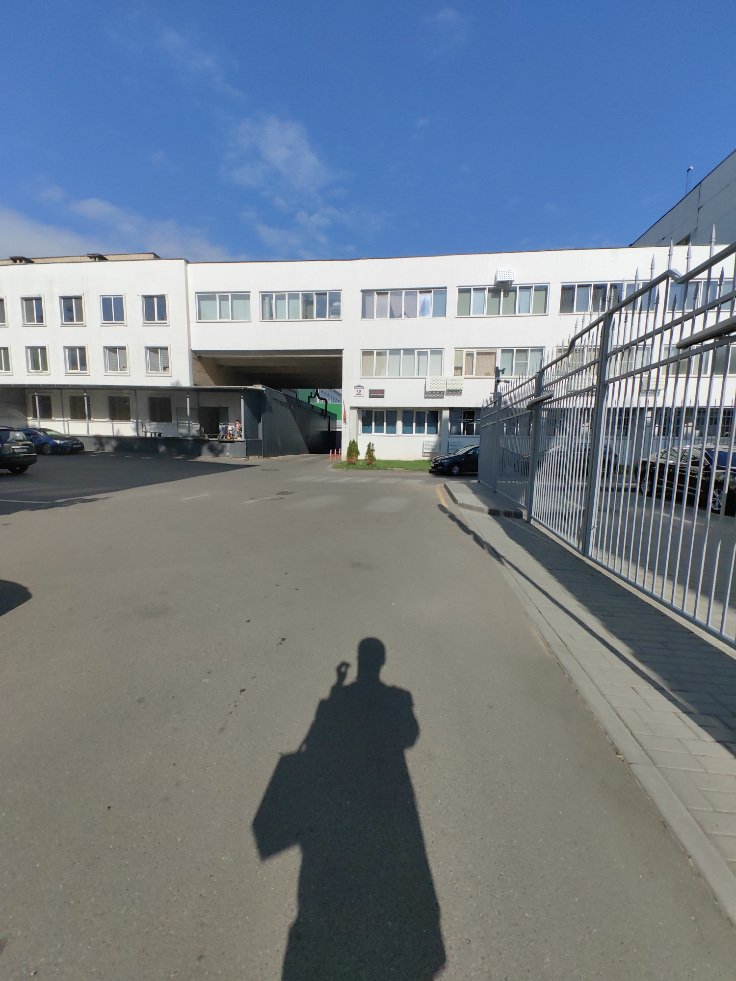 The image shows a grey asphalt pavement area in front of a large, white, multi-story building with many windows. The building appears to be an industrial or commercial facility. To the right of the building, there is a tall, white metal fence. A shadow of a person holding a camera is visible on the pavement in the foreground, indicating the image was taken by a person. Several cars are parked in the vicinity of the building, including one black SUV on the left and other cars behind it. The sky is blue with scattered clouds. The scene is likely an exterior view of a business or factory in Minsk, Belarus.
