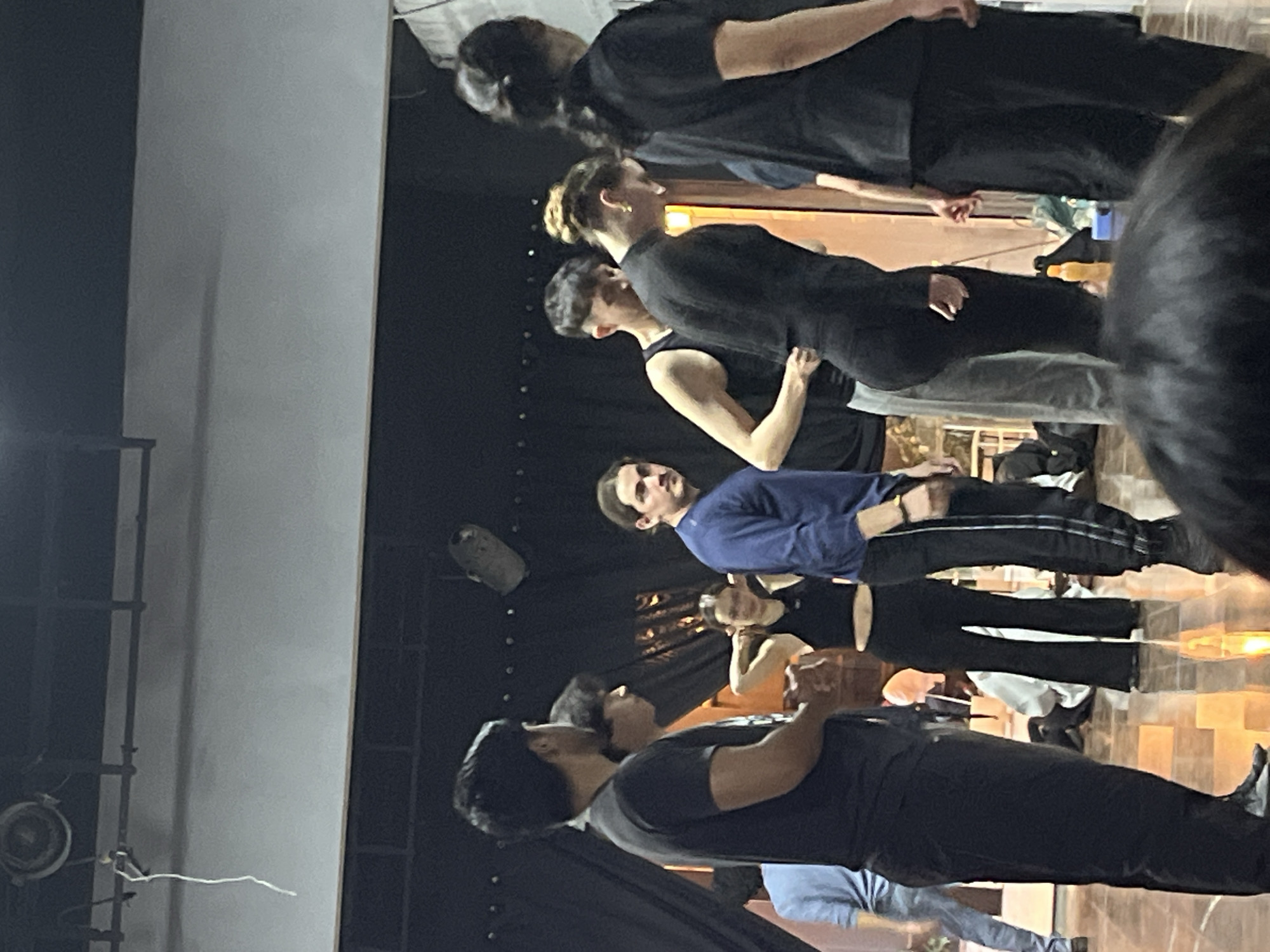The image depicts an indoor scene within the city of San Bernardino Tlaxcalancingo, Mexico, likely a studio or rehearsal space. Approximately ten young adults are present, arranged loosely across the frame. The background consists of dark fabric or curtains, with a lighter-colored wall segment and metallic overhead structures, including a circular light fixture and a cylindrical object, from which a white string hangs. The floor is reflective, showing light glare and partial reflections of individuals.
In the center-left, a male with medium-length dark hair and a beard, wearing a long-sleeved blue shirt and dark track pants, stands looking towards the viewer. To his immediate right, a female with blonde hair tied in a bun, dressed in a form-fitting black long-sleeved top and black pants, is bent forward, looking downwards. Behind her, a male in a black tank top and light gray sweatpants appears to be adjusting his clothing. To the left of the central male, a female in a black sports bra and black wide-leg pants stands with her hands behind her head, also looking towards the viewer. On the far left, two males in black short-sleeved shirts are partially visible, one in profile. A person in a light blue shirt and jeans is partially seen further left. On the far right, a female with dark hair tied back, wearing a black short-sleeved top, faces away from the camera. A blurred head with dark hair is present in the immediate foreground, indicating the photographer's position behind another person. A yellow water bottle and various bags or clothing items are discernible on the floor in the mid-background. The individuals are standing in varied postures, suggesting a break, warm-up, or period of transition within an activity.