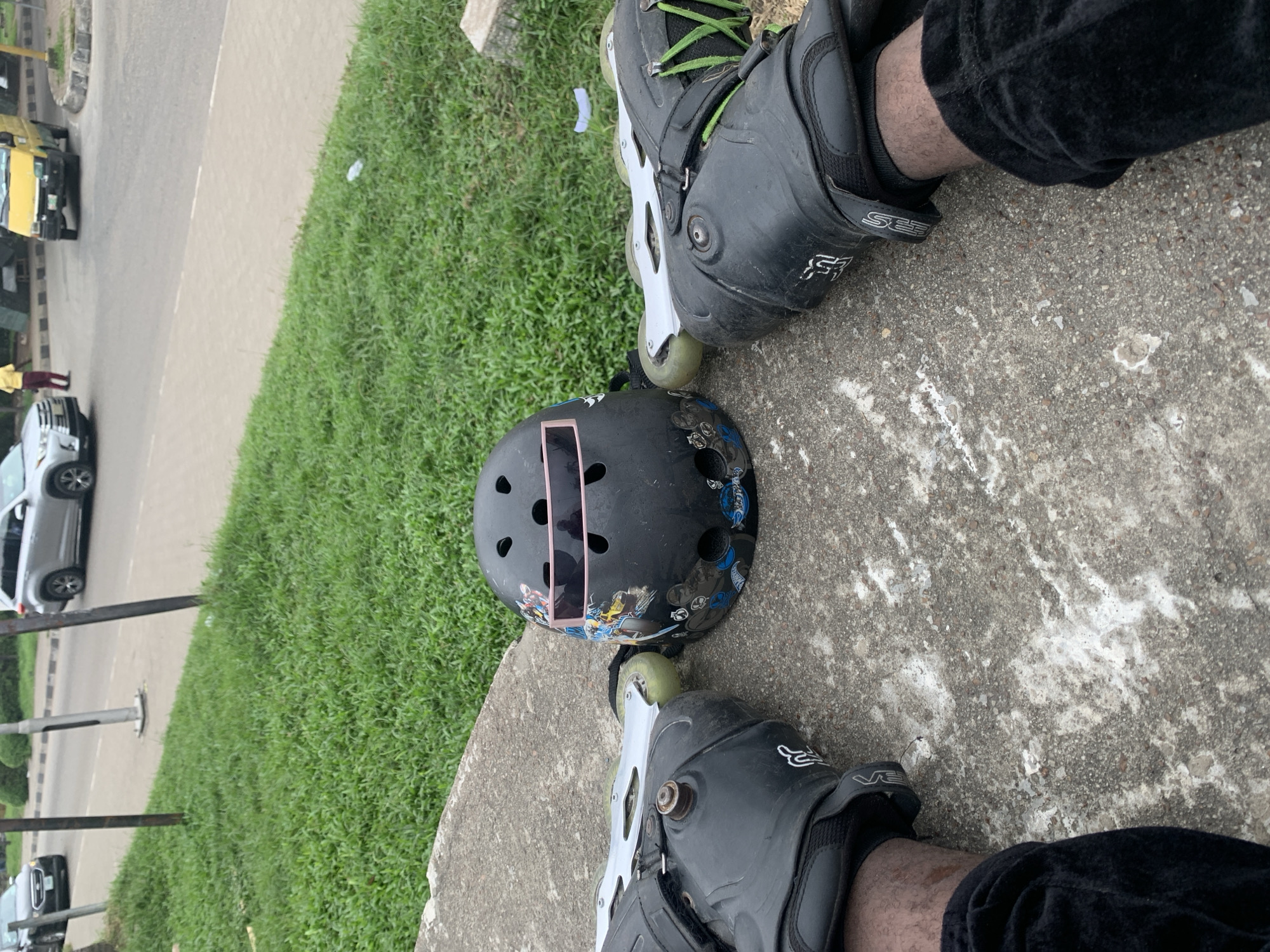 In Iganmu, Nigeria, a black helmet with a pink visor adorned with stickers sits on a concrete surface next to a pair of black inline skates. The skates are positioned on the feet of a person wearing black pants and black socks. The scene overlooks a grassy area and a road where a silver SUV and a yellow bus are visible, along with a person in a yellow shirt and red pants standing near the bus.