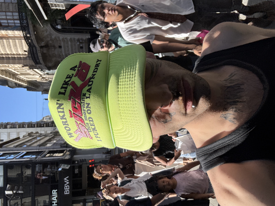 A daylight, close-up photograph captures a male individual in the foreground, largely obscured by a prominent neon green mesh trucker hat, against the backdrop of a busy street scene in Buenos Aires, Argentina. The hat features the text "WORKIN' LIKE A SICKO / THIS MORNING I PUKED ON LAUNDRY" in stylized black and red lettering. The subject's lower face is visible, revealing smudged dark marks or makeup under both eyes, a small metal ring piercing in the right nostril, and a metal stud labret piercing below the lower lip. Tattoos resembling dark brushstrokes or patterns are visible on his throat and neck. He wears a dark, ribbed tank top.
The background depicts a crowded urban street lined with multi-story, ornate European-style buildings, characteristic of central Buenos Aires architecture. A "BBVA" bank sign is clearly visible on the facade of a building on the left side, along with a partial "HAIR" sign above it. Numerous pedestrians populate the street, including a woman adjusting her hair, a young child in a pink top, and a man in a white shirt and patterned tie on the right side. The scene is brightly illuminated by direct sunlight under a clear blue sky.