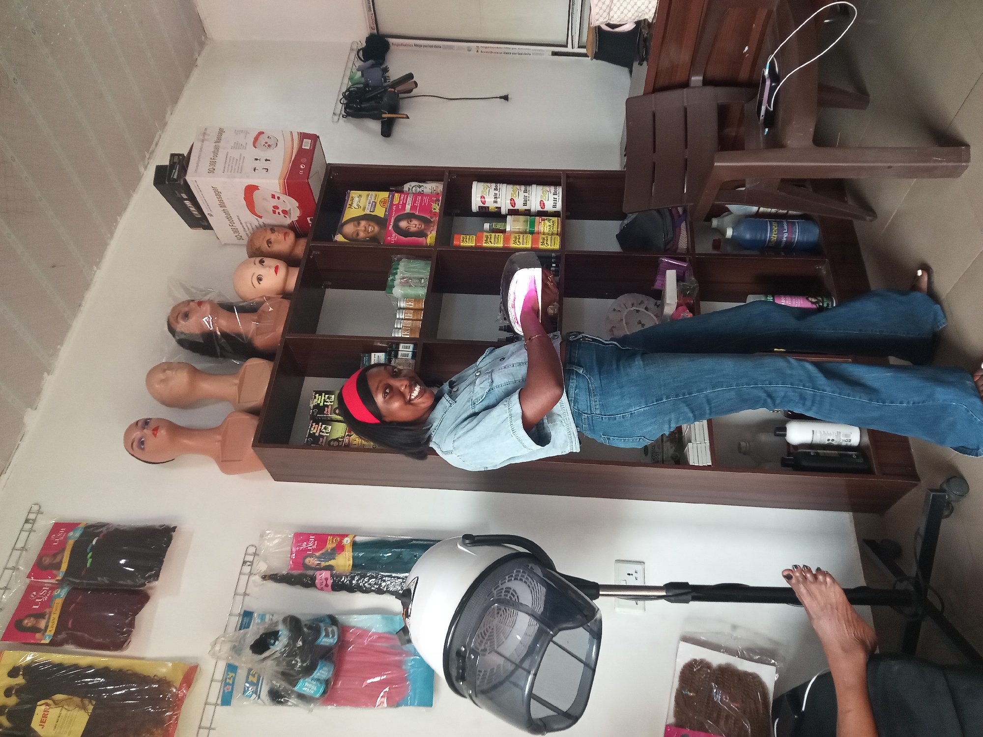 A smiling young woman, dressed in a light blue denim shirt and blue jeans, stands in what appears to be a hair salon or beauty supply shop in Sabon Tasha, Nigeria. She is holding a smartphone in her right hand, looking directly at the camera with a happy expression. A red headband adorns her dark hair.
Behind the woman is a multi-tiered wooden shelving unit, fully stocked with various hair care products and beauty items. Visible product names on the shelves include "Moro Growth", "Hair Boo", and "treen ting Lotio". Cardboard boxes for "SQ-368 Footbath Massager" and "SQ-168 Footbath Massager" are also stacked on top of the shelves. To the left of the shelves, several mannequin heads are displayed on a white counter, some with hair.
Below the shelving unit and on the adjacent white wall, various packaged hair extensions and wigs are hung on wire racks, with labels such as "LUSH", "JERR", and "WOOL" visible on some. A white and grey stand-up hair dryer with a dark hood is positioned to the right of the woman, plugged into a wall socket. A dark brown wooden chair is partially visible in the upper right background. The setting conveys a lively indoor space, characteristic of a local beauty business.